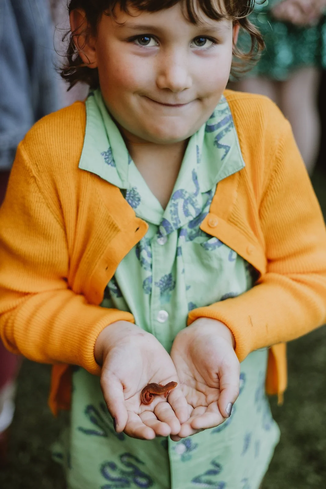 A young boy with curly hair and light skin holding a small wiggly worm in his cupped hands, smiling slightly.