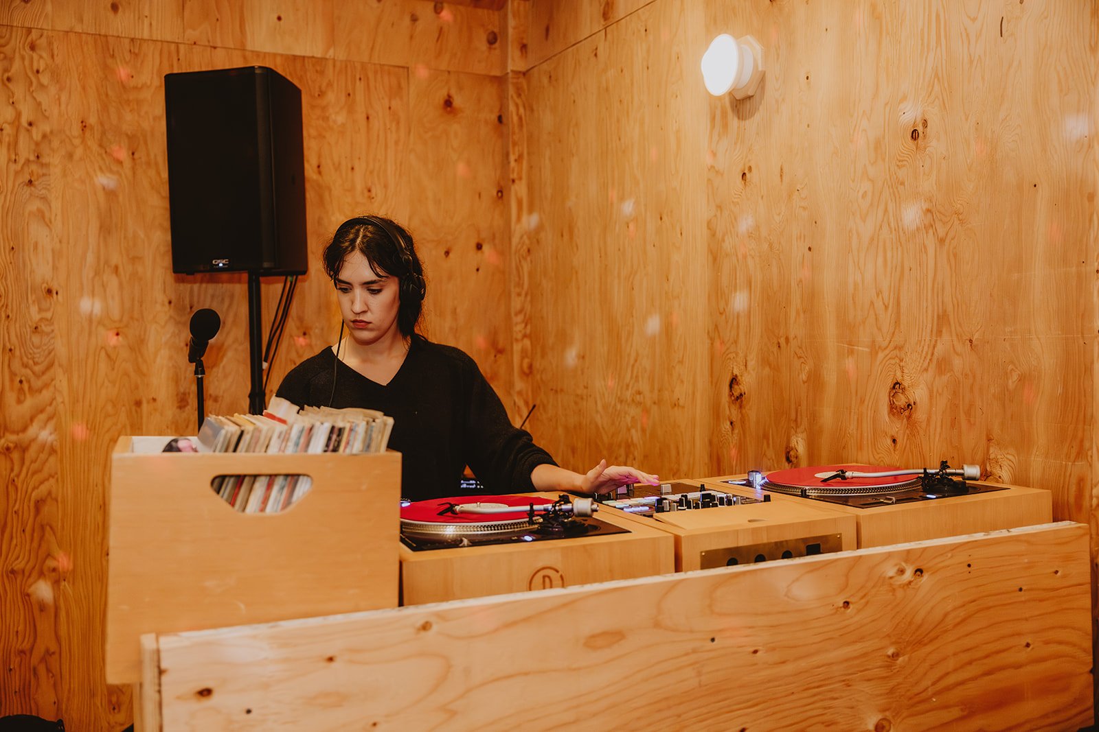 A woman DJ in a wooden booth with vinyl records and turntables, with a large speaker and microphone.