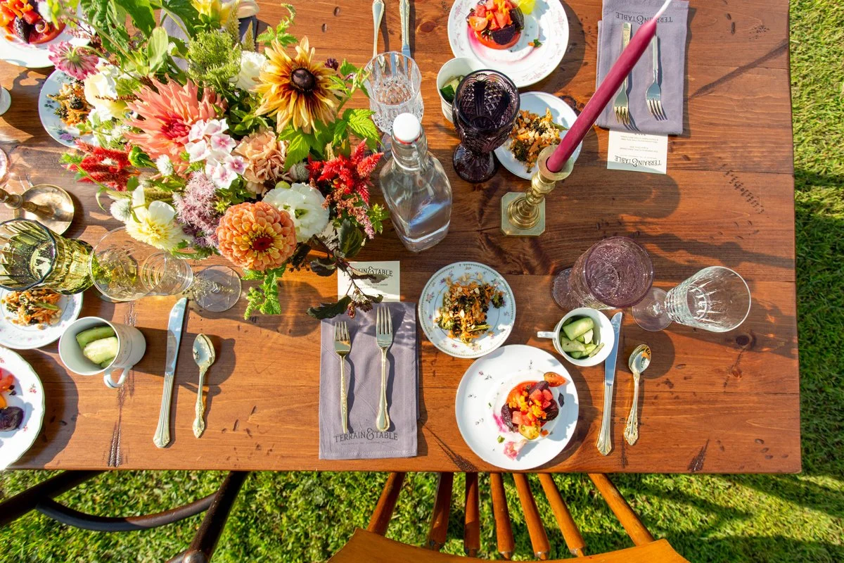 A beautifully arranged outdoor dinner table set with floral centerpiece, plates of salad, cutlery, glasses, a pink candle in a holder, and bowls with cucumbers, on a sunny day on green grass.