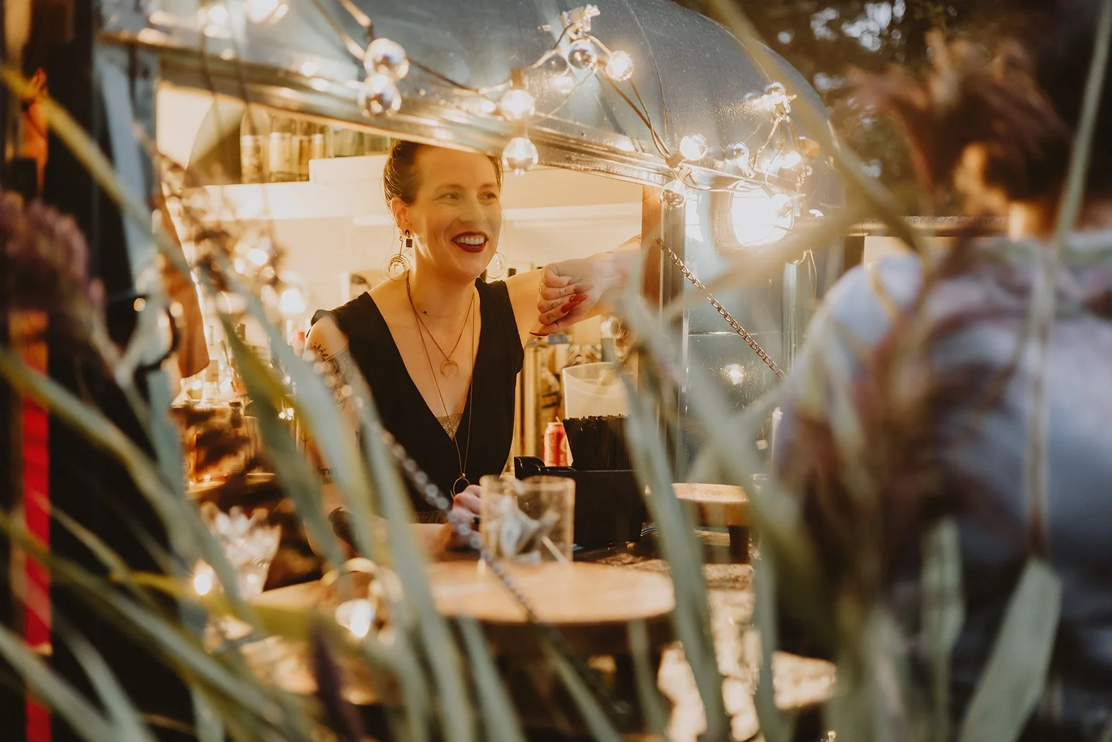 A woman smiling and greeting someone at a booth or bar, seen through a decorative frame with string lights and plants, at night.