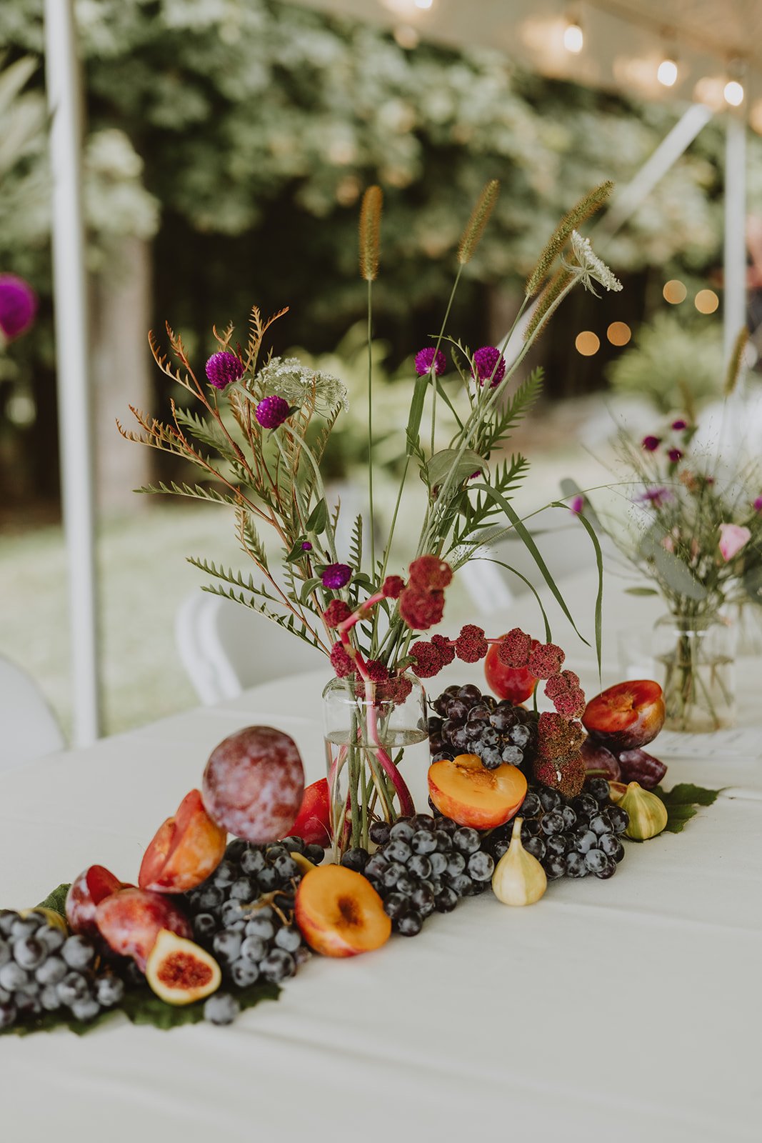 A floral centerpiece with purple, red, and white flowers and greenery in a glass vase, surrounded by grapes, figs, peaches, and garlic on a white tablecloth, set outdoors with blurred background.
