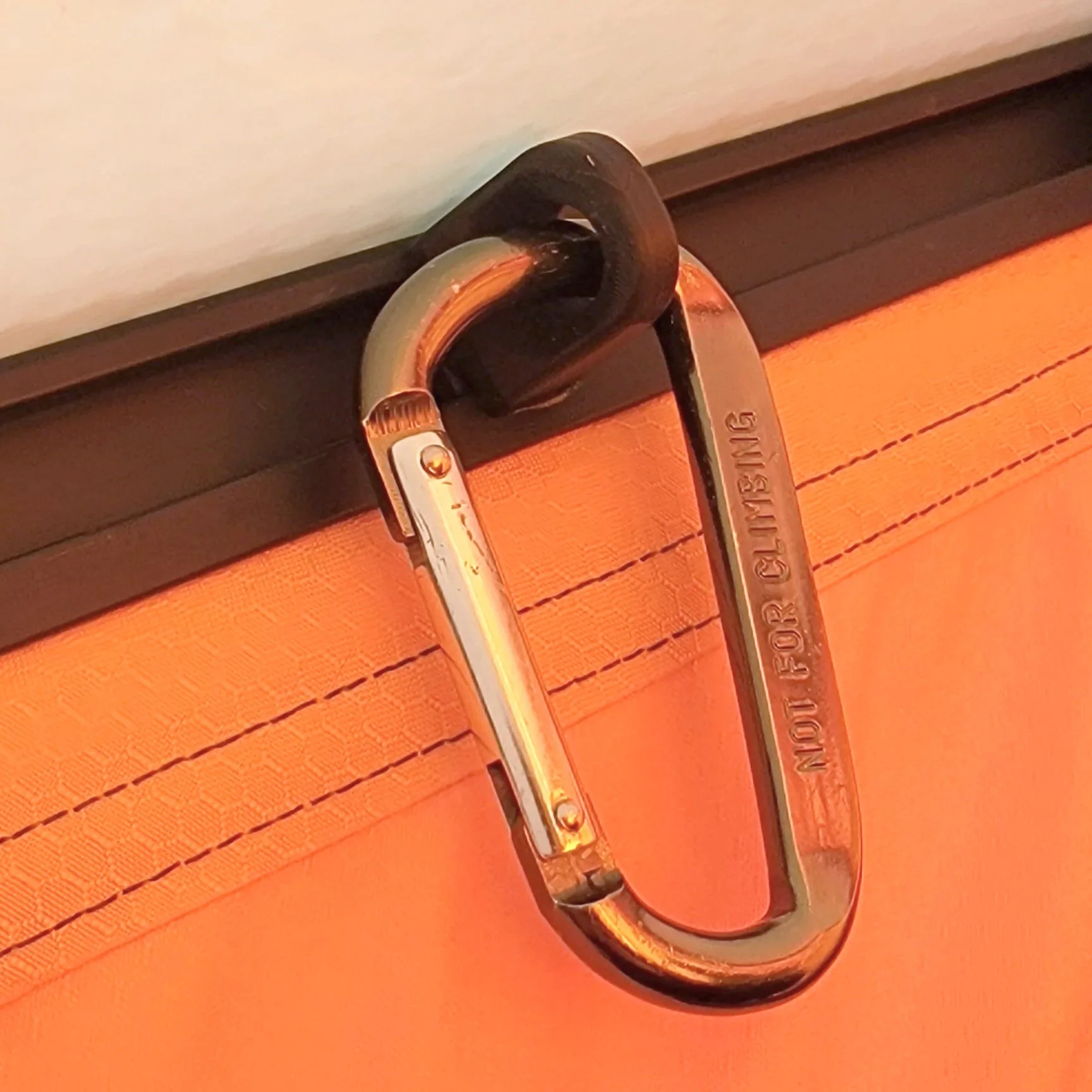 Close-up of an orange backpack with a black strap and a metal carabiner attached to it. The carabiner has the words 'MOLLE FOR CLIMBING' engraved on it.