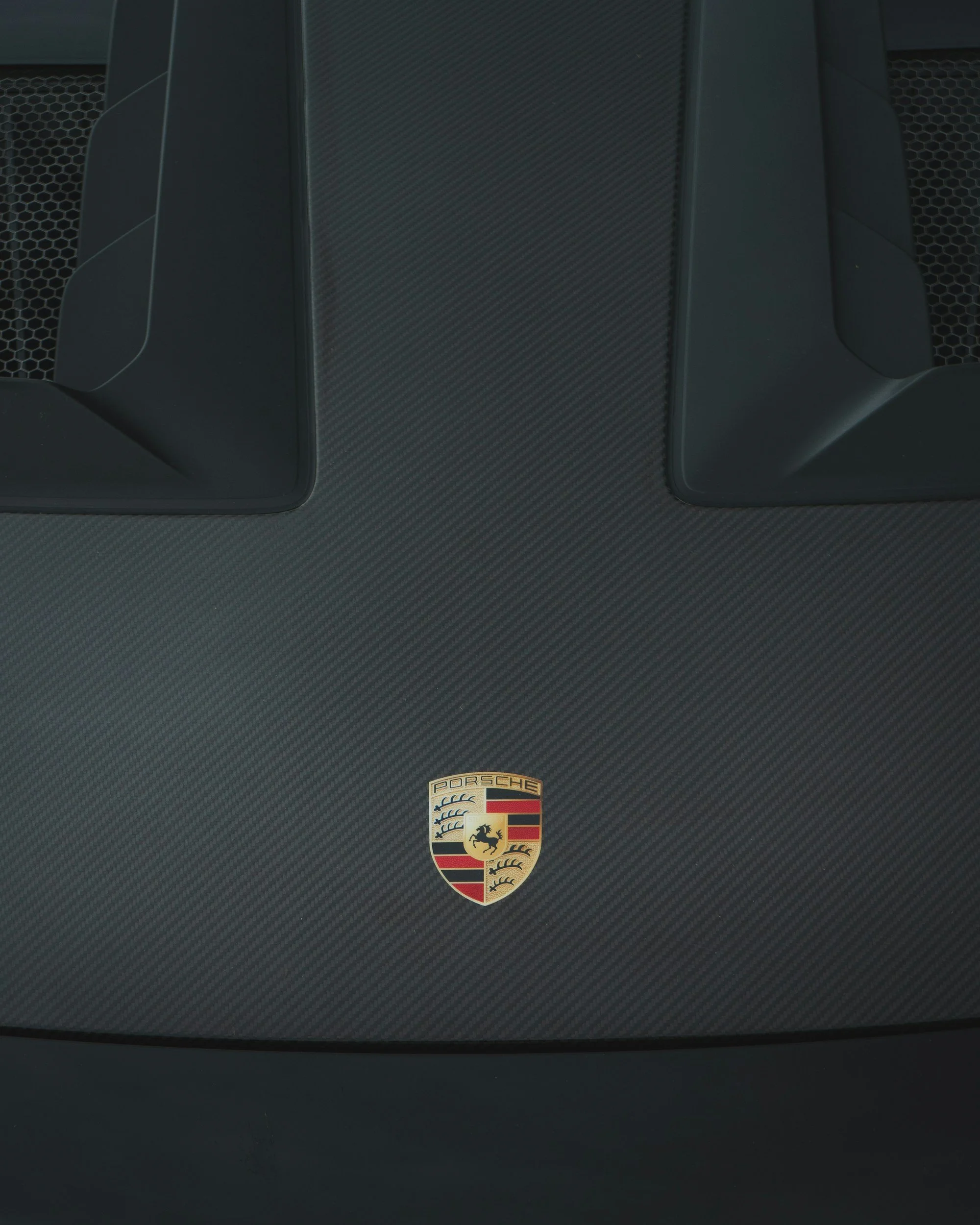 Close-up of a Porsche emblem on a dark, textured car hood with two black vents above it.