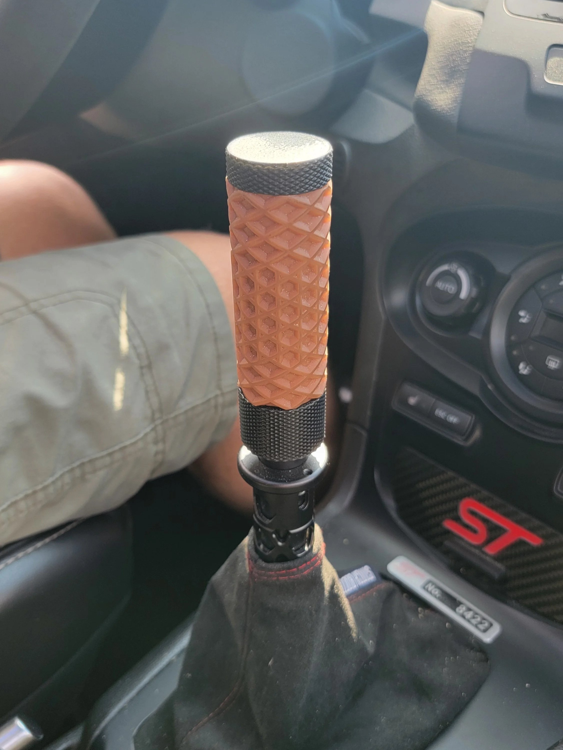 Close-up of a car's gear shift with a custom grip, featuring a textured, orange material and black accents, in the interior of a vehicle with manual transmission.