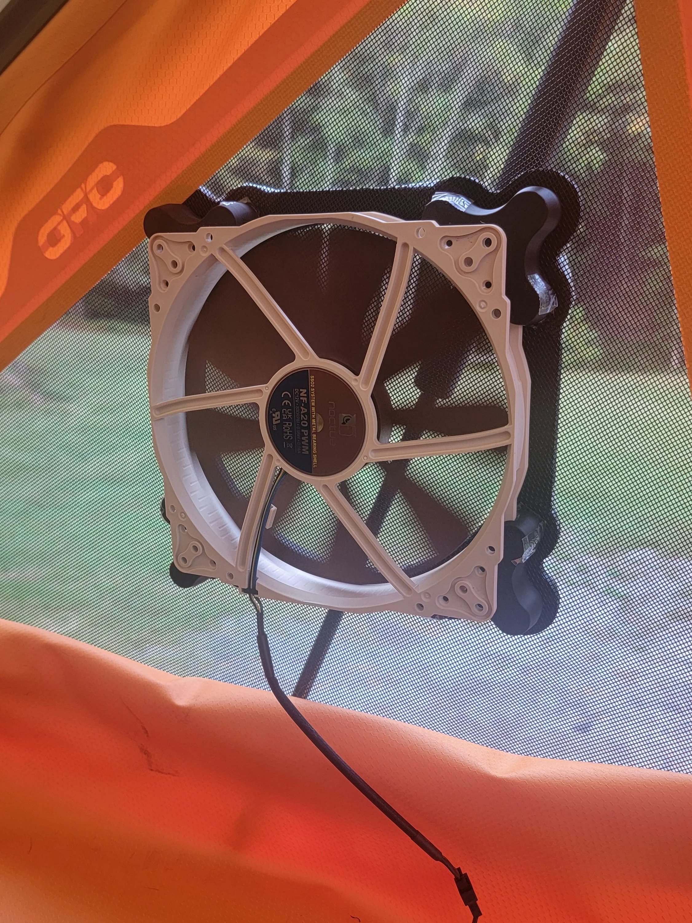 A computer cooling fan mounted inside a mesh enclosure, with an orange fabric at the bottom and part of a canopy or tent in the background.