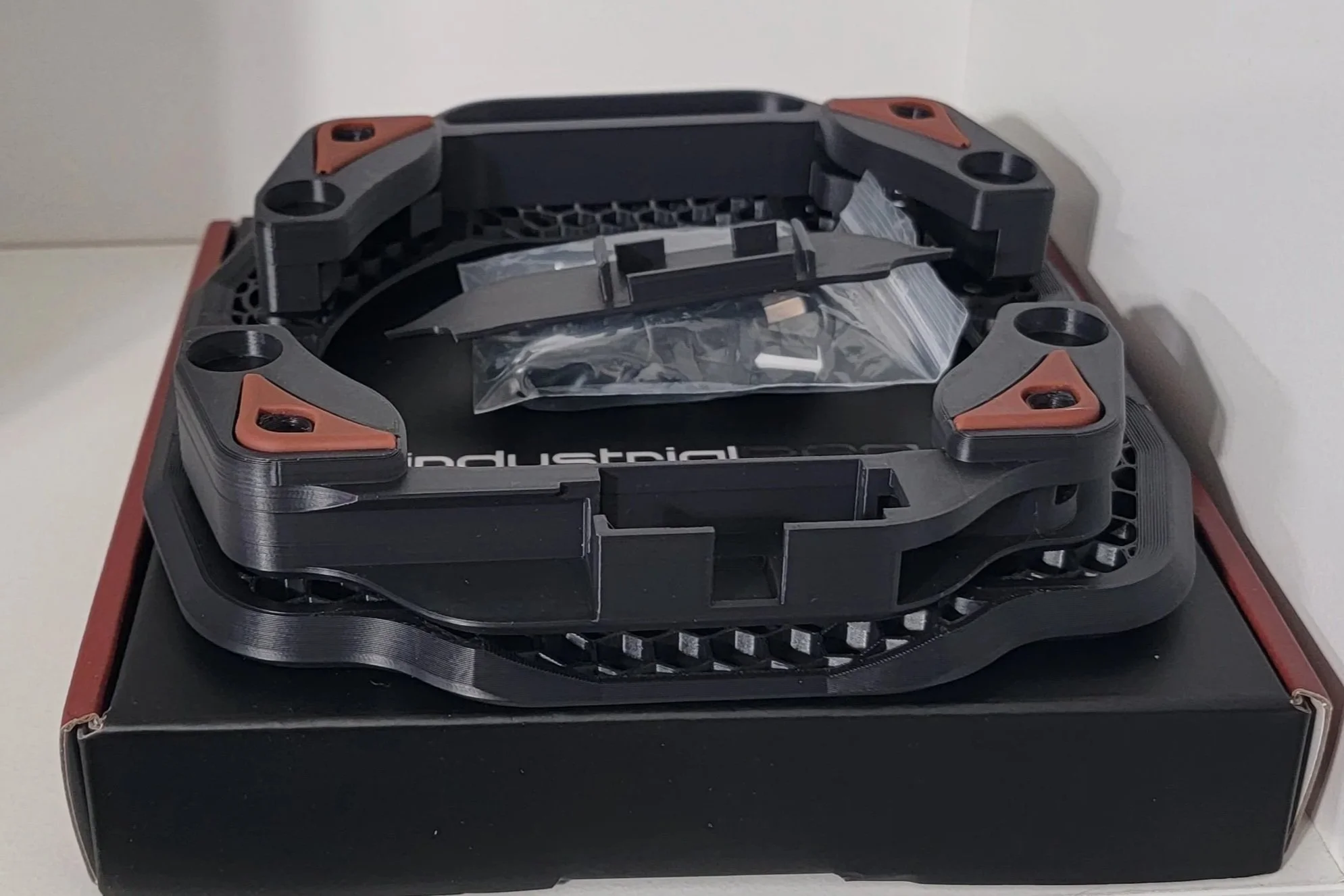 A 3D-printed race drone frame placed on top of a black box, with a small plastic bag and some screws inside. The frame has a circular central area with four supporting arms and orange accents at the corners.
