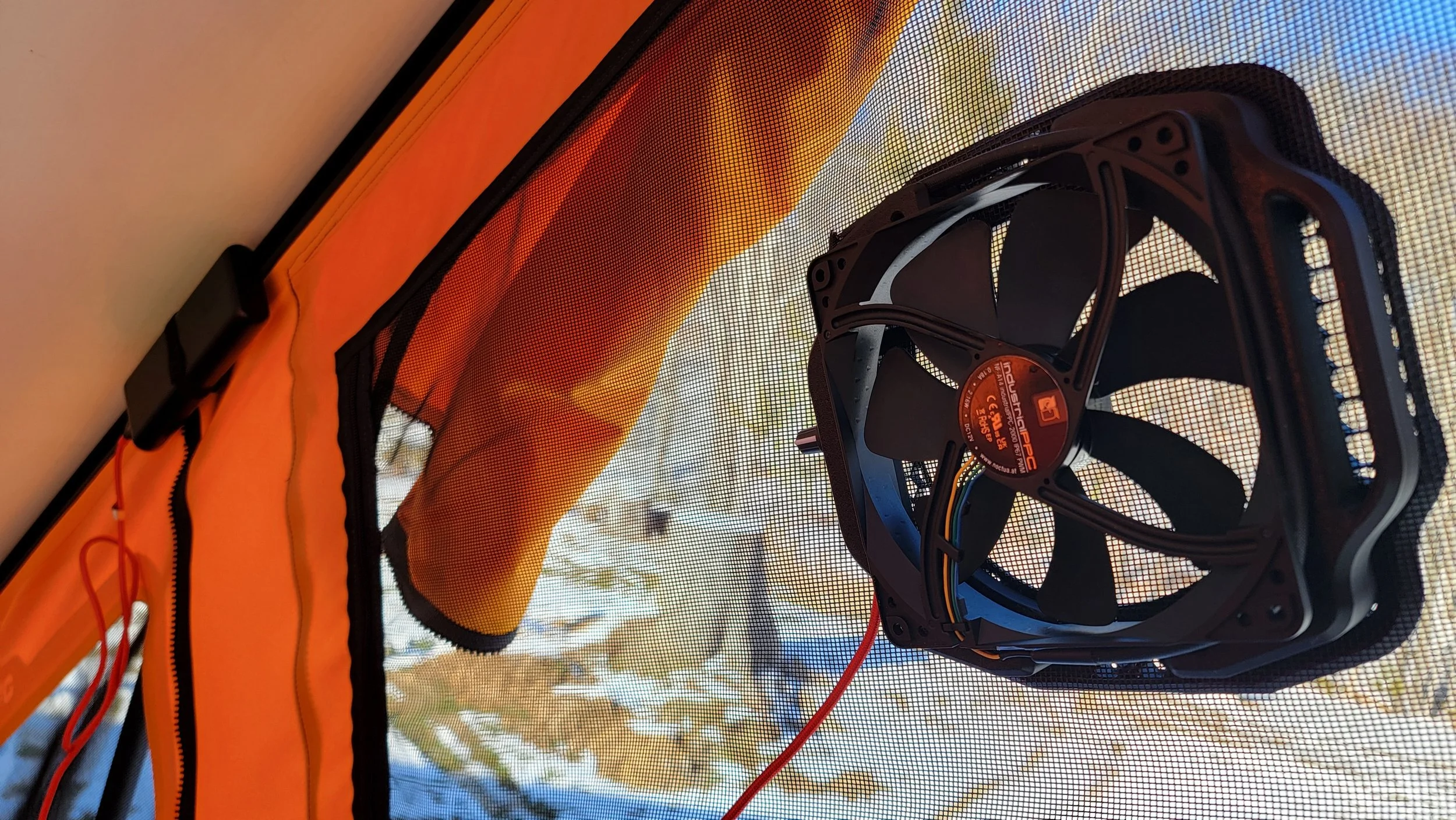 Close-up of a computer or cooling fan mounted inside a tent or fabric enclosure with orange and black material. The fan has a label with technical specifications and is connected by wires.