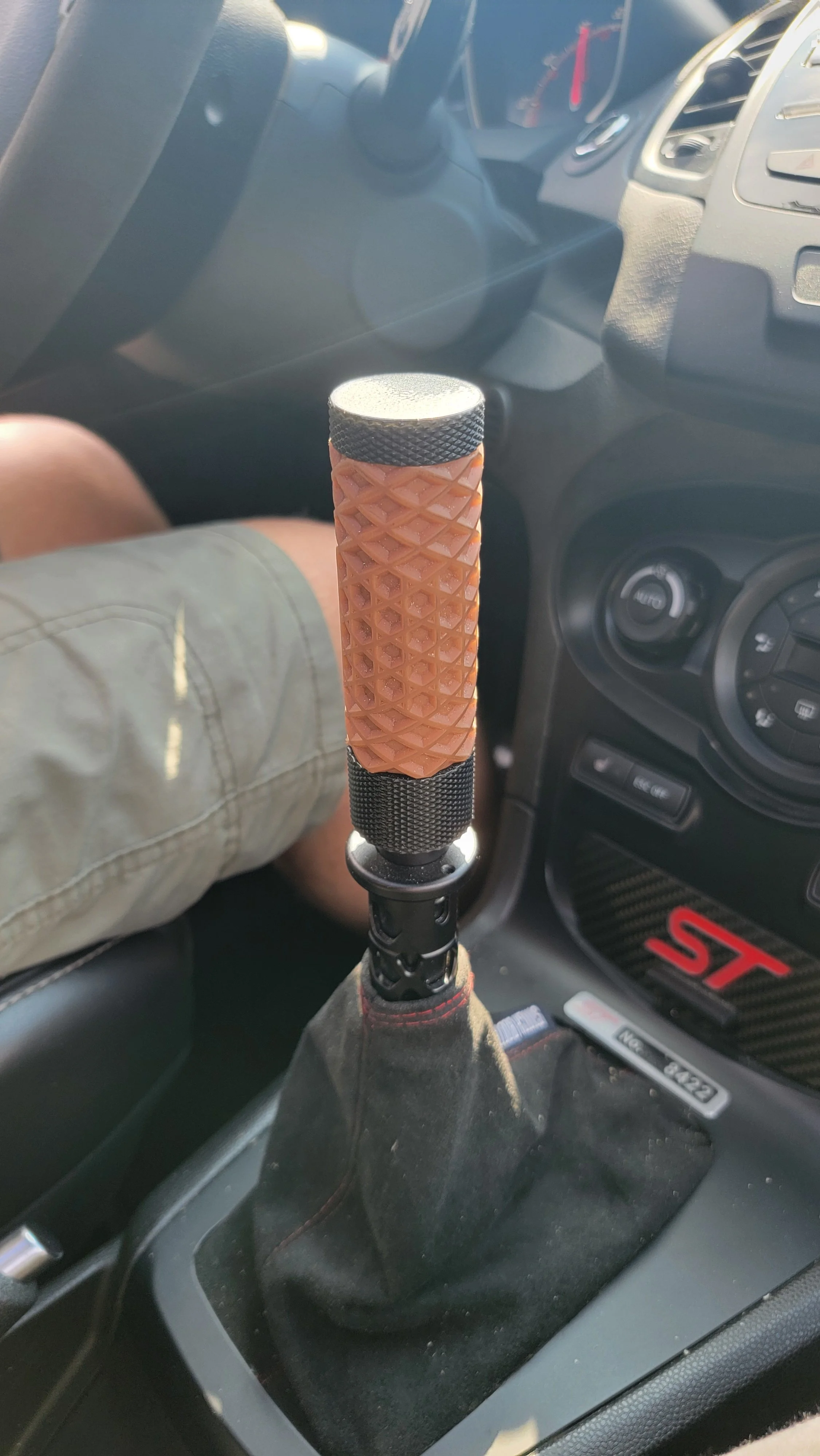 Close-up of a car's gear shift with a custom brown textured knob inside a vehicle with a black interior and a ST logo on the dashboard.