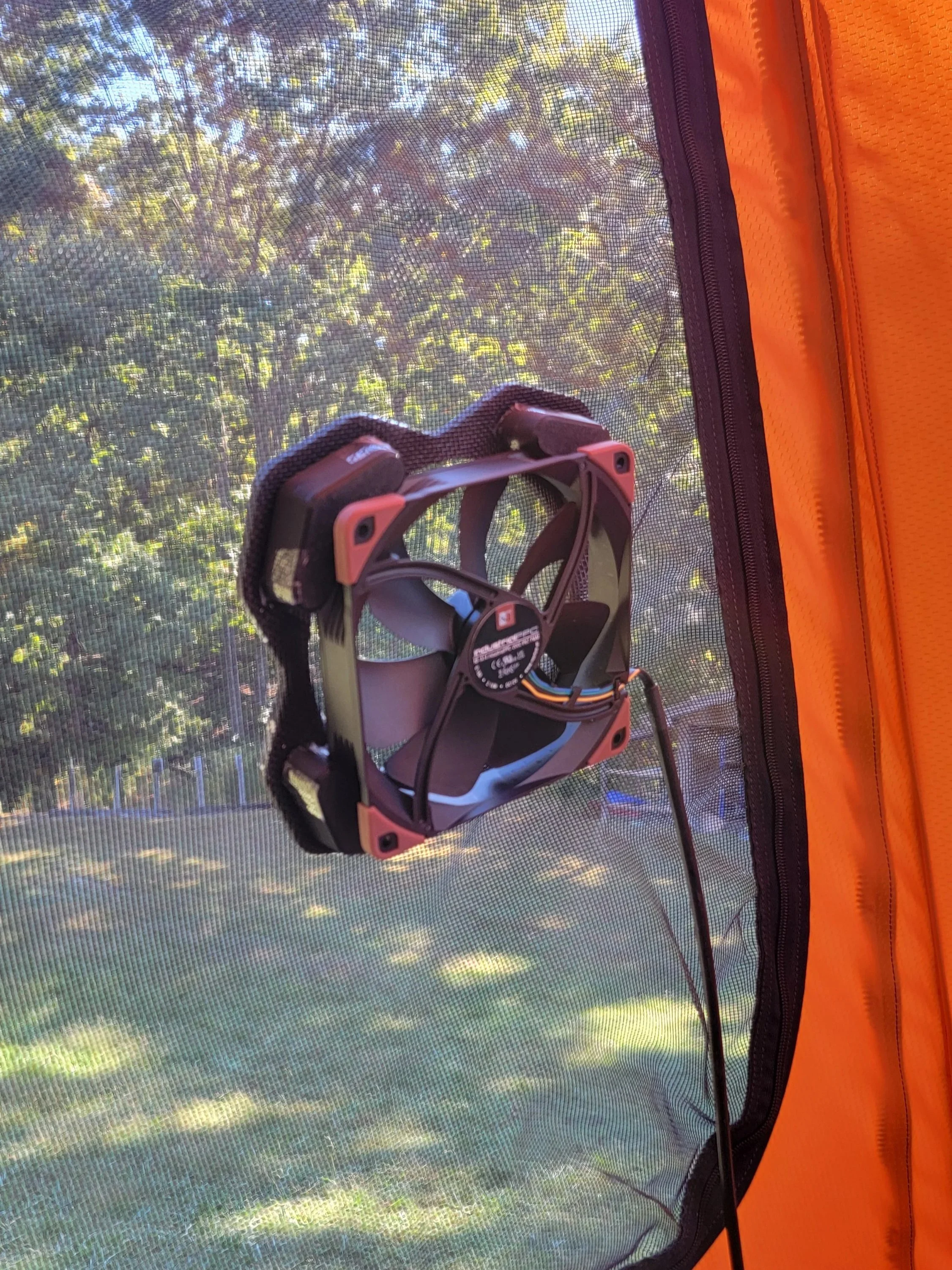 A small black and red portable fan attached to a mesh window screen, outside of an orange tent, with trees and blue sky in the background.
