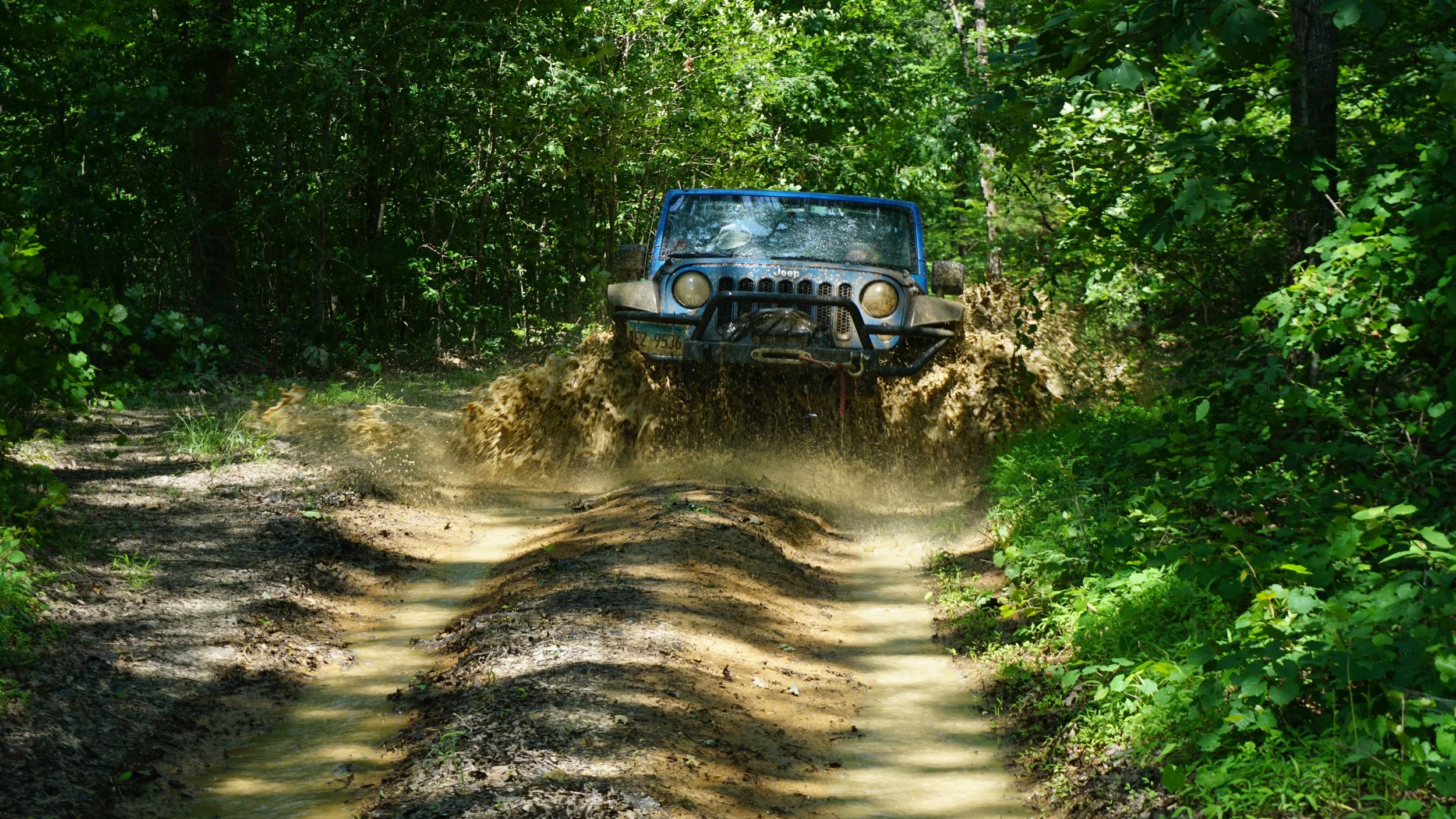 A blue Jeep driving through a muddy forest trail, splashing mud and water while surrounded by green trees and foliage.