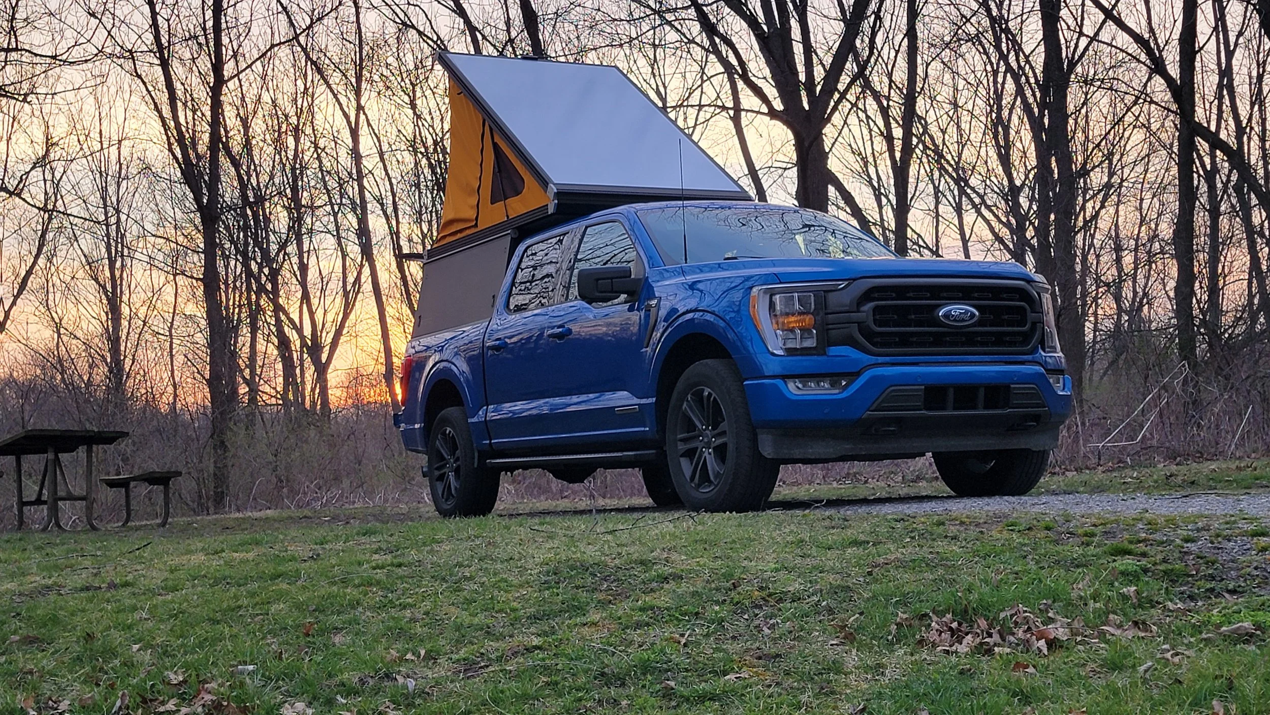 A blue Ford F150 pickup truck parked outdoors during sunset with a roof-mounted tent. There are leafless trees in the background and two picnic tables to the left.