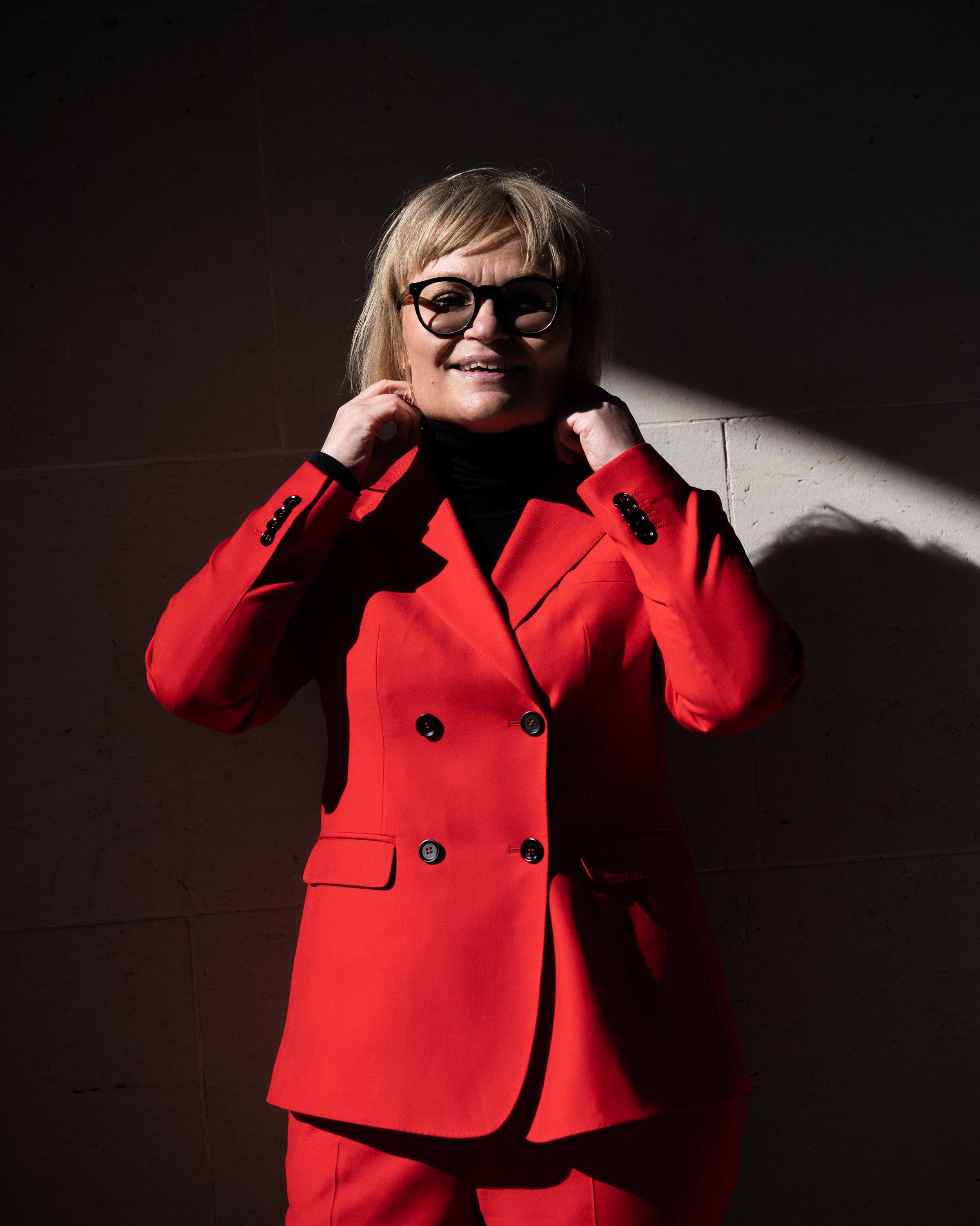 Kvindelig person, iført rød frakke med store sorte knapper, står mod en mørk væg med skygge, har blond hår og sorte briller, smiler og justerer halsklædet.