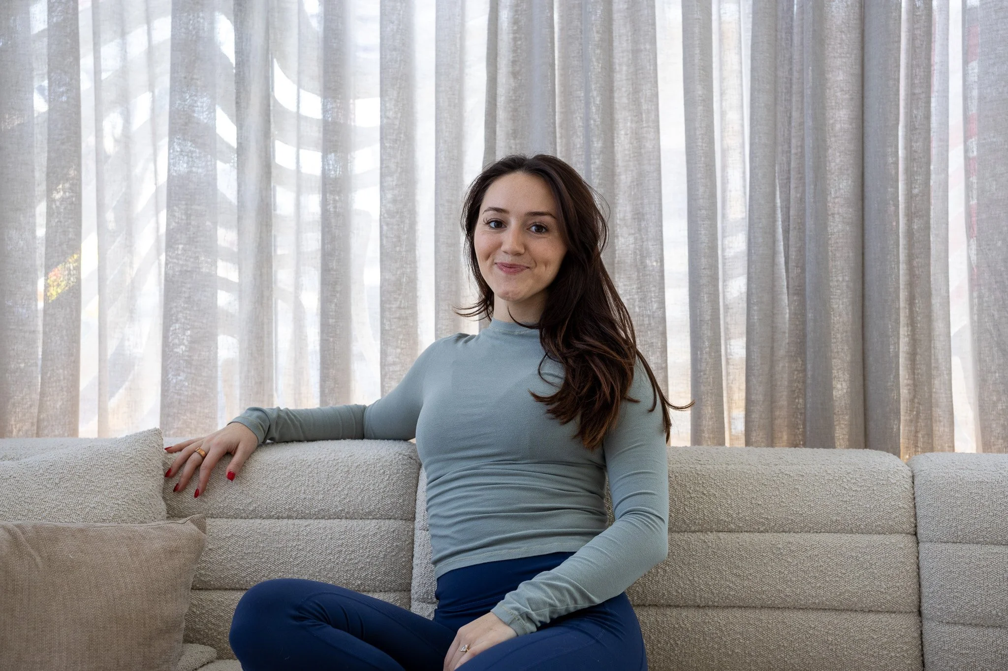 A woman with long brown hair sitting on a beige sofa in front of sheer curtains, smiling slightly at the camera.