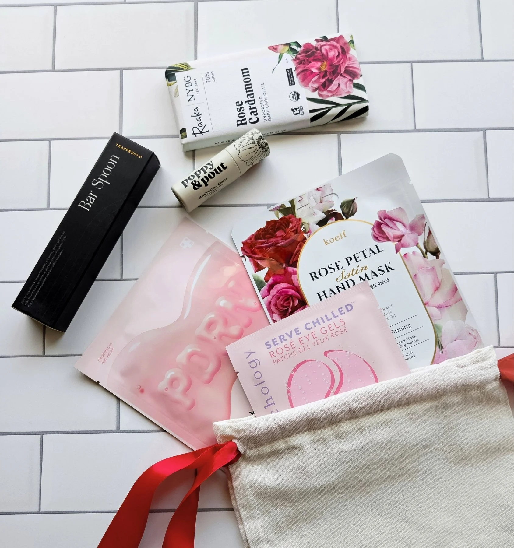 Curated Valentine’s gift bag featuring a sheet mask, rose eye gels, chocolate bar, lip balm, and hand mask in a cotton drawstring bag.