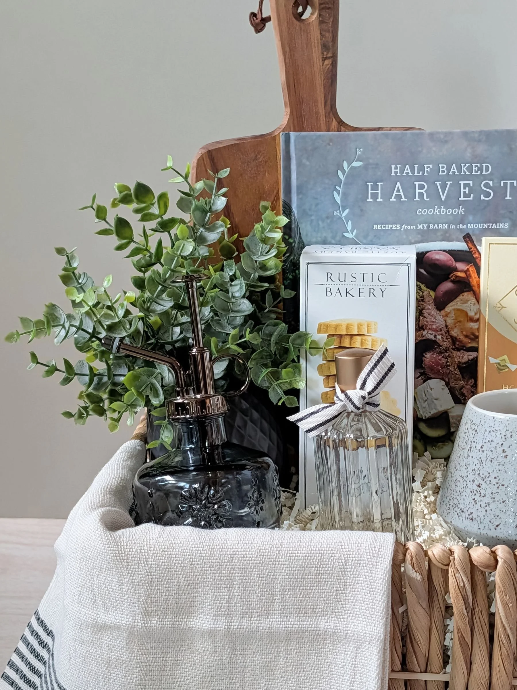 Warm, neutral‑toned gift basket with cookbook, gourmet treats, plant mister, Turkish towel, artisan mug, and faux floral accents