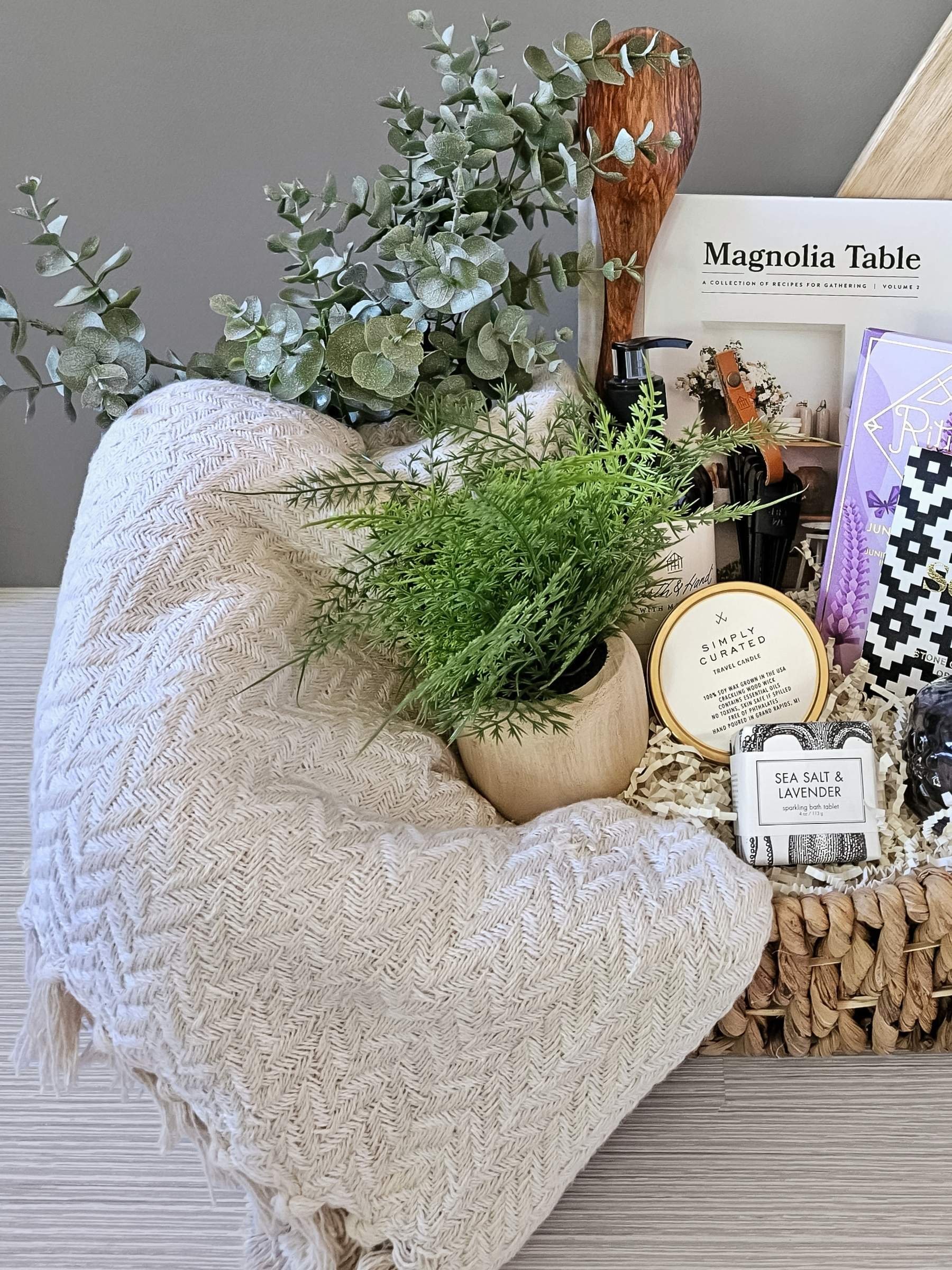 Elegant housewarming gift basket with a textured throw, artisan chocolates, bath bomb, candle tin, serving board, and stylish home essentials.
