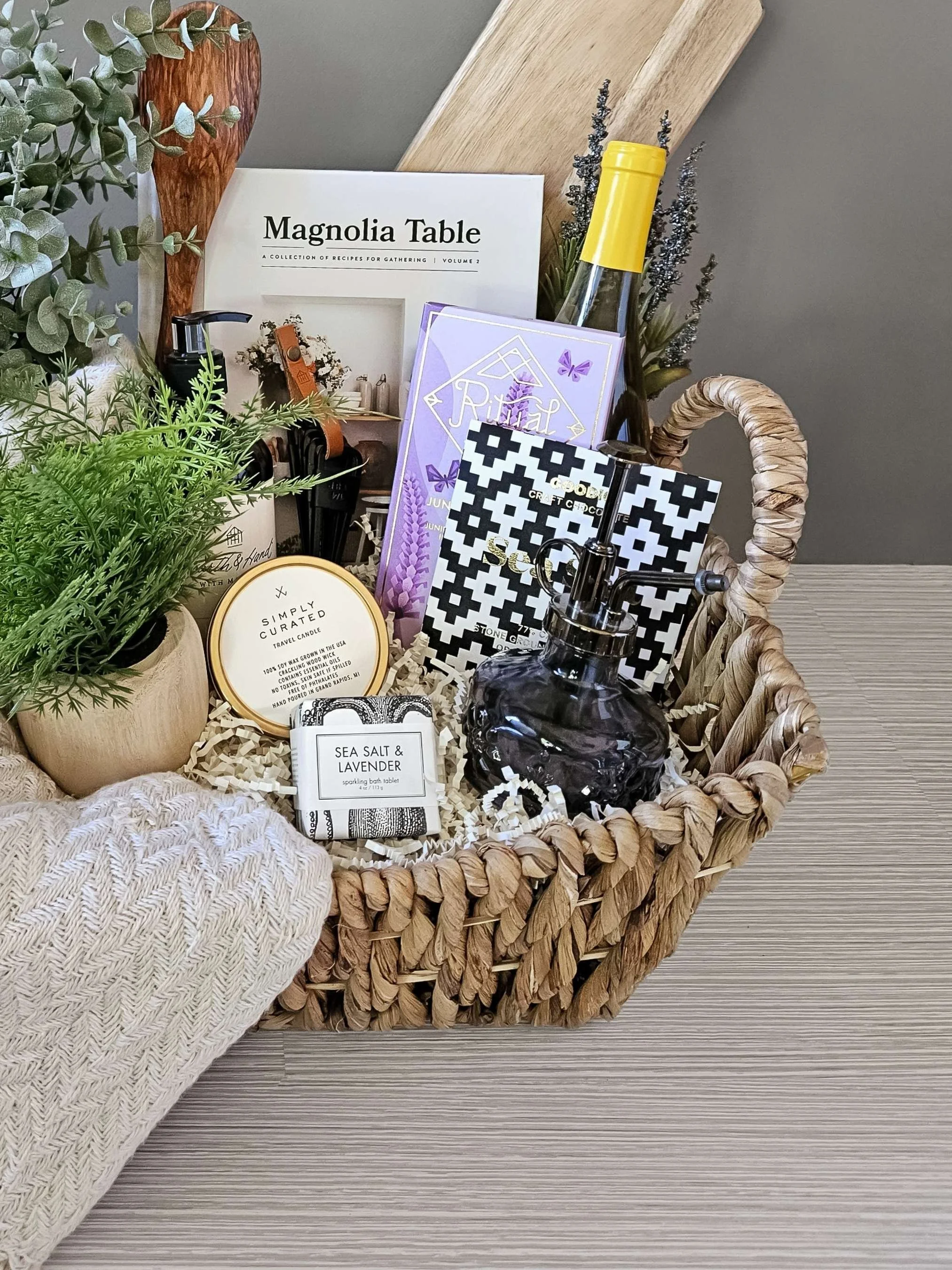 Curated home‑inspired gift box featuring a cozy throw, gourmet sweets, bath bomb, candle, plant mister, and refined serving pieces.