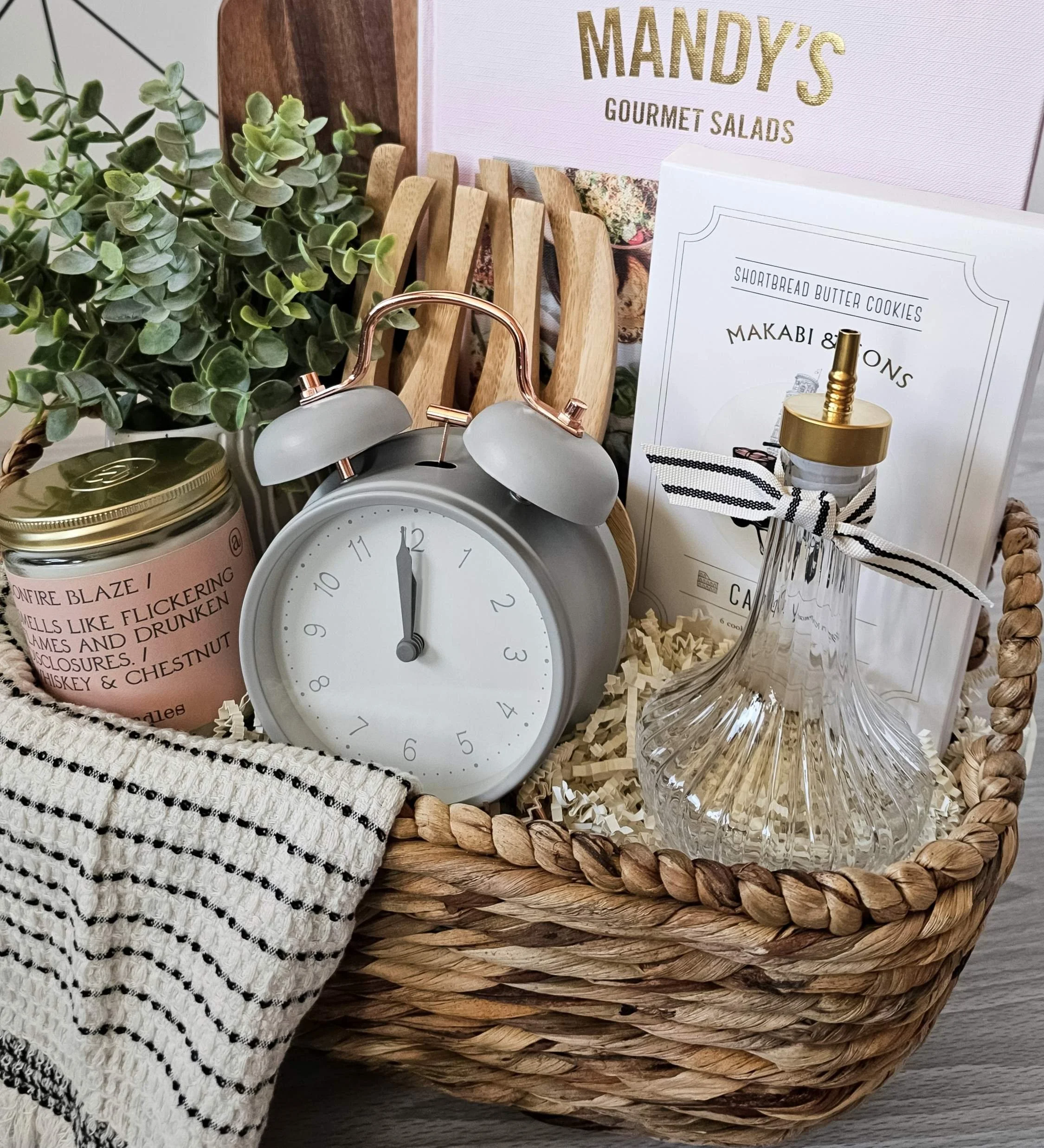 Light and feminine spring gift basket featuring cookbook, wood board, candle, salad claws, cookies, and a textured woven hand towel.