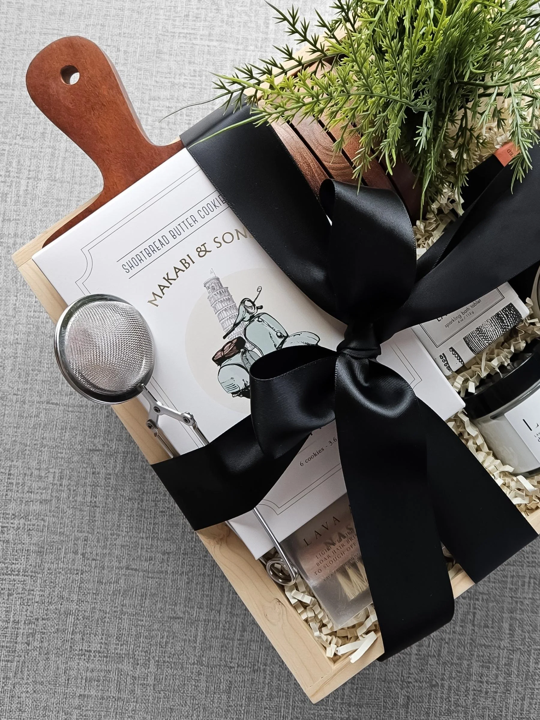 Curated gift with lemon poppy shortbread cookies, a wood serving board, a boar and lava brush, and a stainless tea strainer arranged in a wooden box.