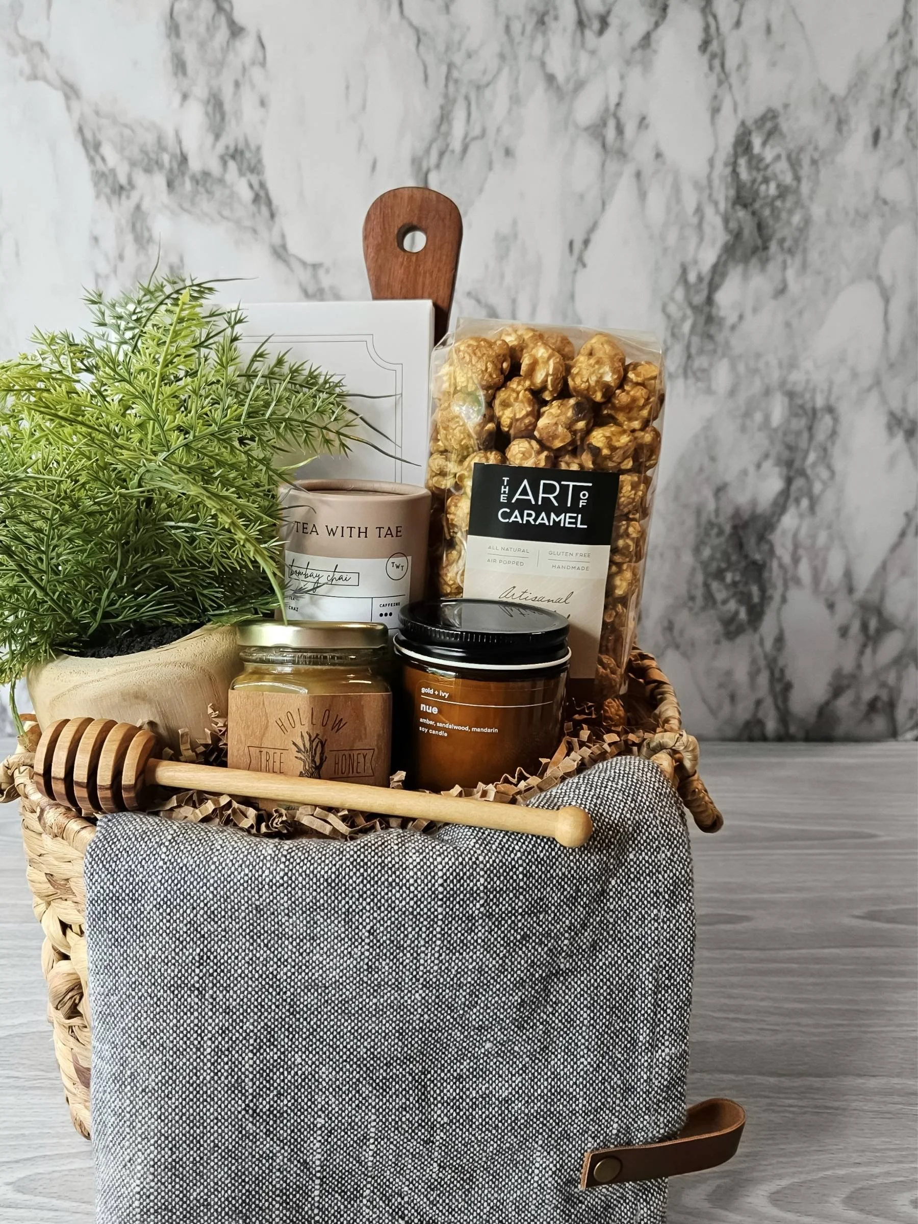 A housewarming gift basket with chai tea, pistachio shortbread cookies, small batch honey, gourmet popcorn, candle, wood board, and a denim tea towel