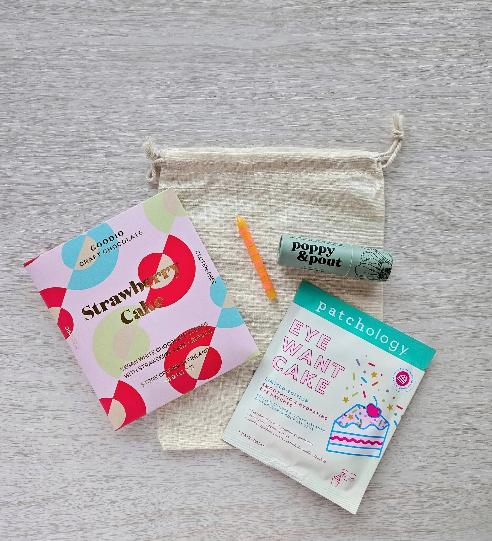 Birthday gift bundle with colorful candle, Eye Want Cake eye gels, strawberry cake chocolate bar, and lip balm arranged on a cotton bag.