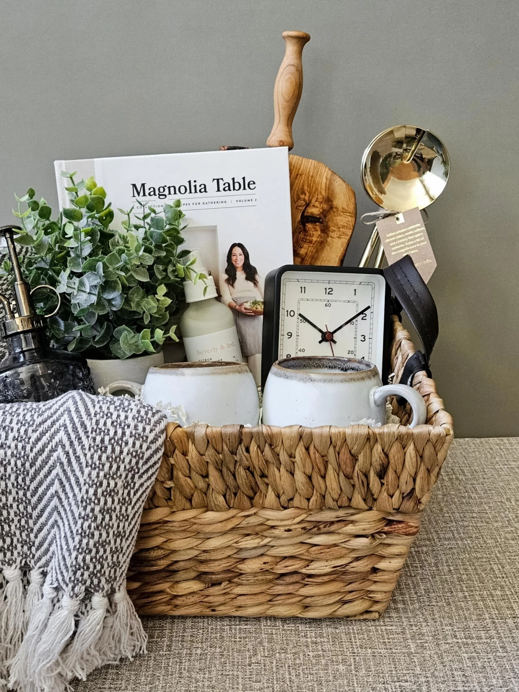 Elegant cozy home gift basket with a plush throw, mugs, serving board, linen spray, plant mister, and refined decorative accents.