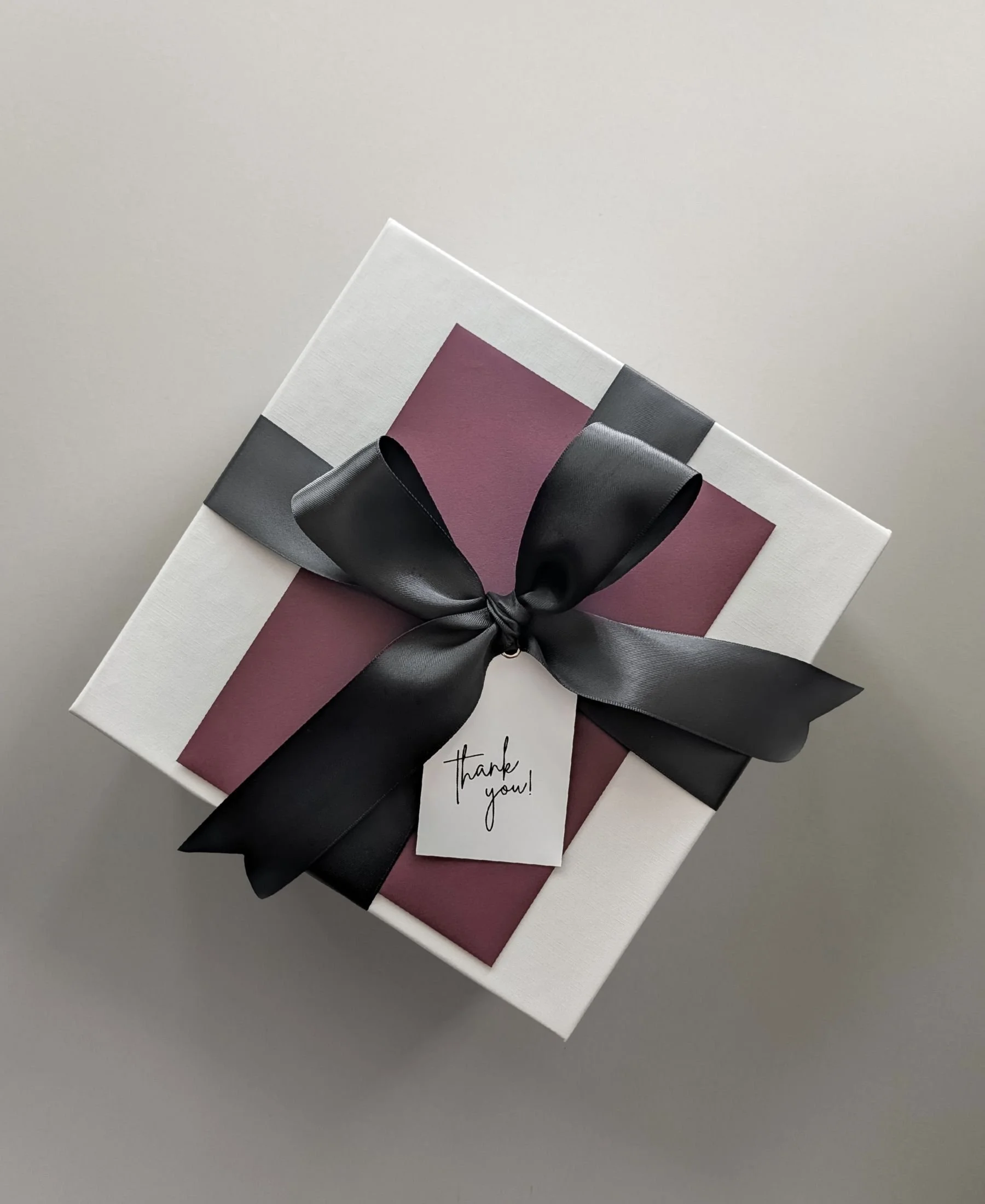 Light grey linen gift box with black double satin ribbon, thank‑you tag, and a personalized notecard tucked inside a burgundy envelope for an elevated presentation.