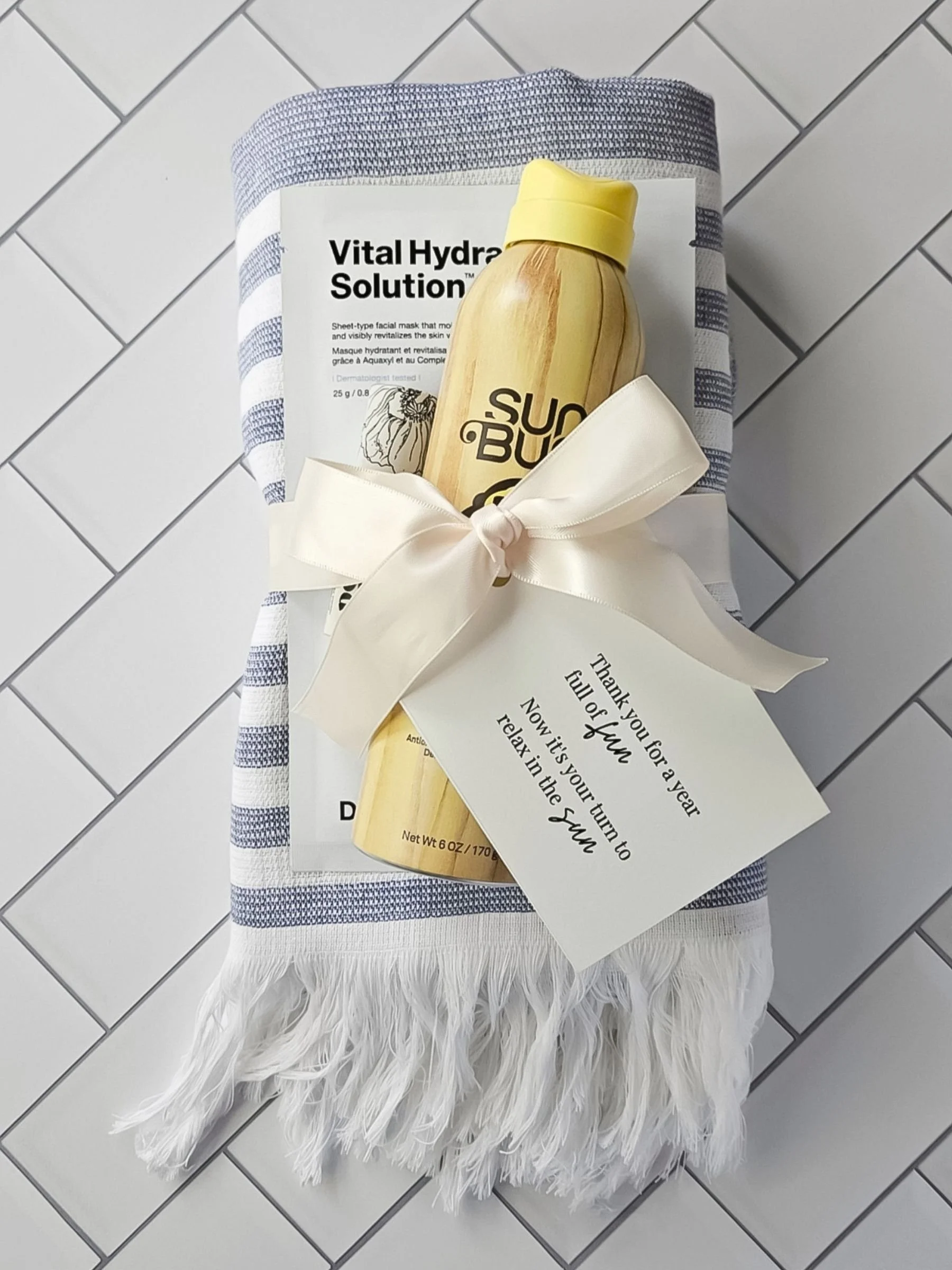 A folded striped Turkish pool towel tied with a ribbon alongside Sun Bum sunscreen and a hydrating face mask, arranged as a summer teacher gift with a printed tag.