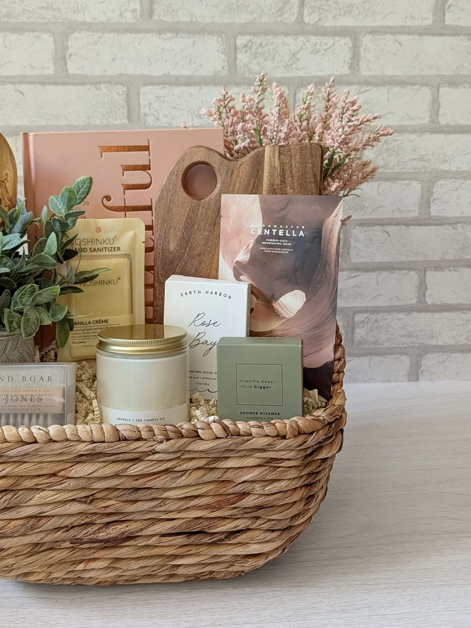 Chic wellness inspired gift basket with a shower steamer, facial cleanser, Centella sheet mask, wood serving board, and candle.