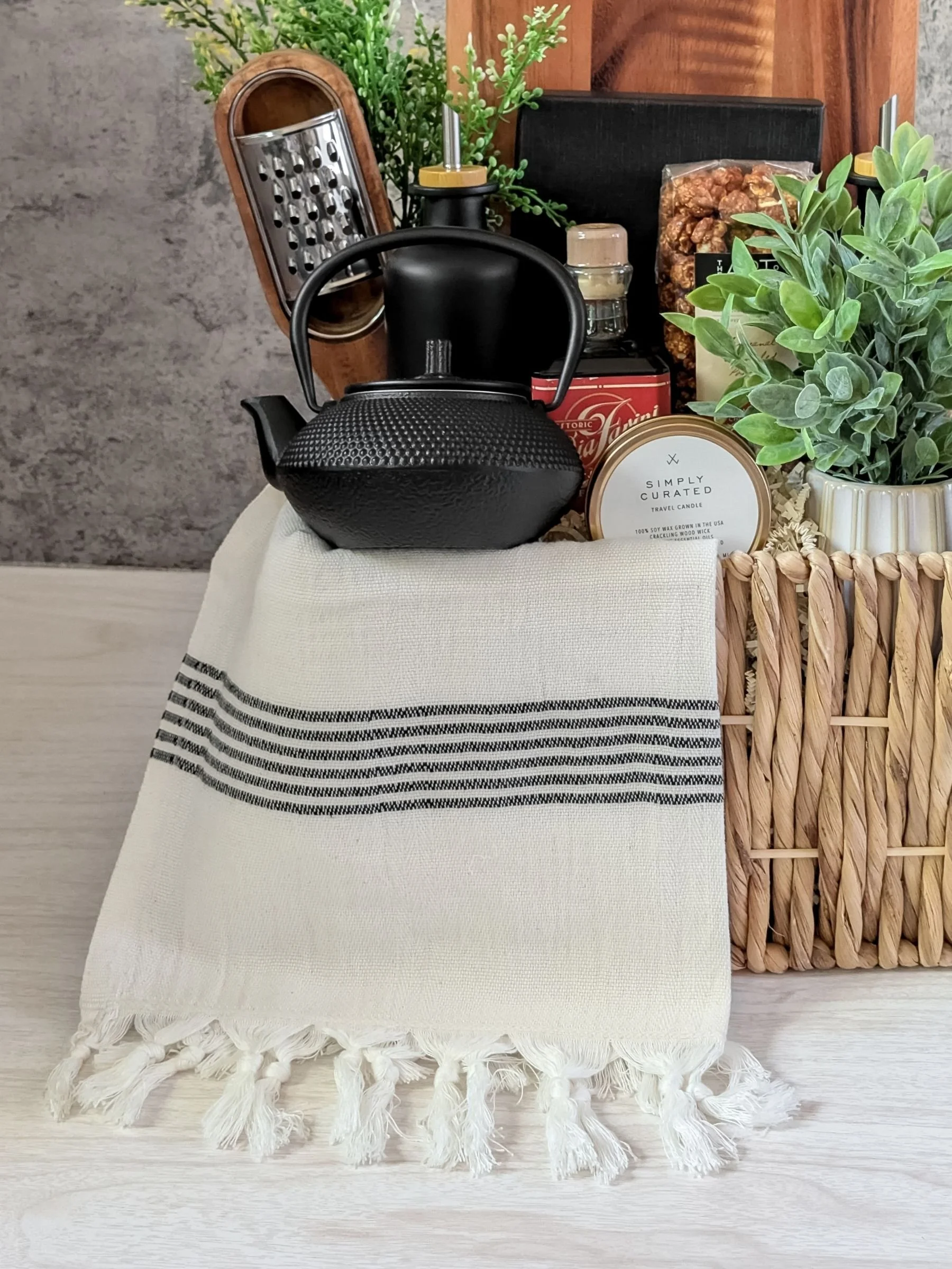 Gourmet modern gift basket featuring a cast iron black teapot, large wood board, luxury wood charcuterie knives, Turkish hand towel, and artisanal gourmet items.