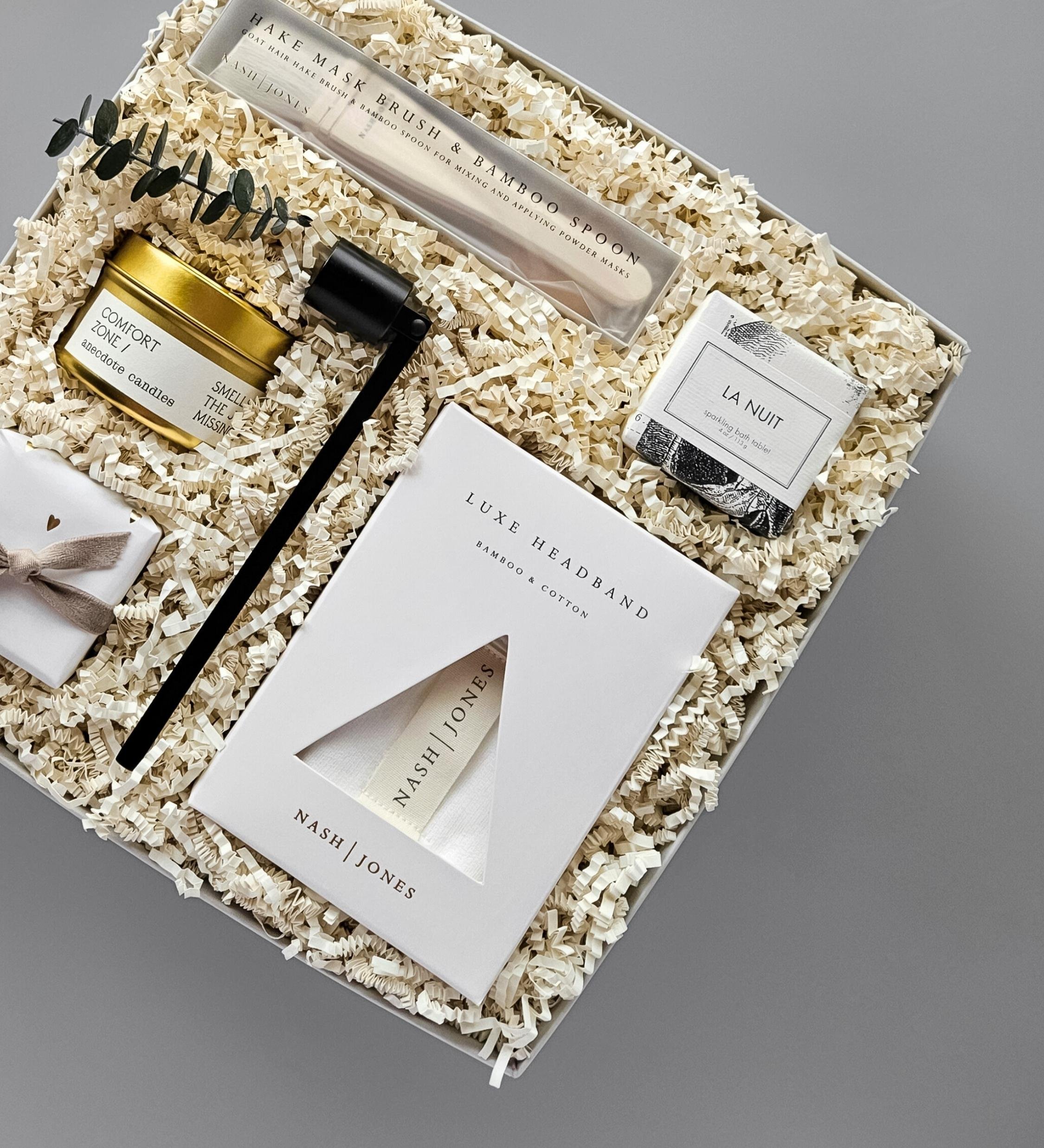 Homebody pampering gift set with headband, bath bomb, face mask, candle, bamboo spoon, mask brush, and matte black candle snuffer.