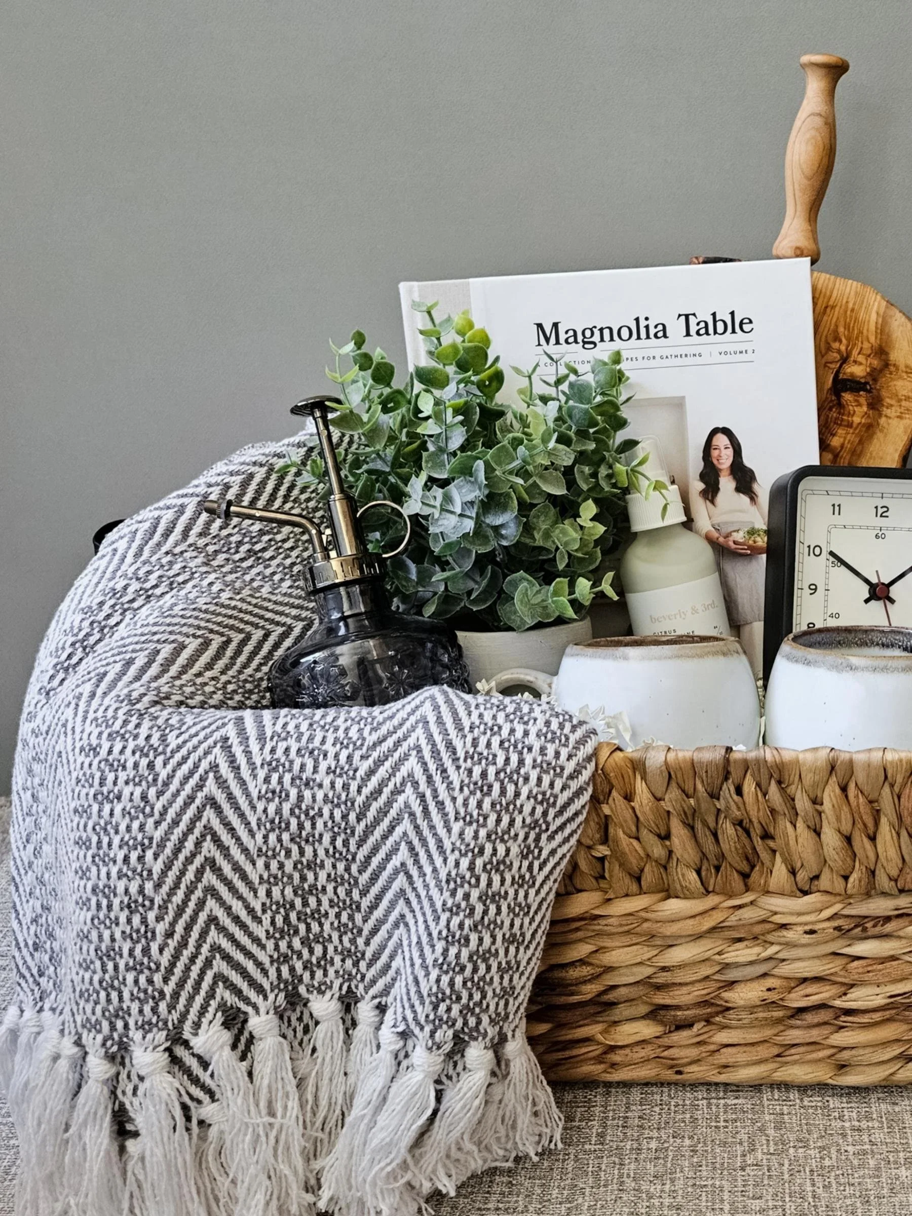 Cozy home gift basket with a herringbone throw, ceramic mugs, serving board, plant mister, and elegant home accents.