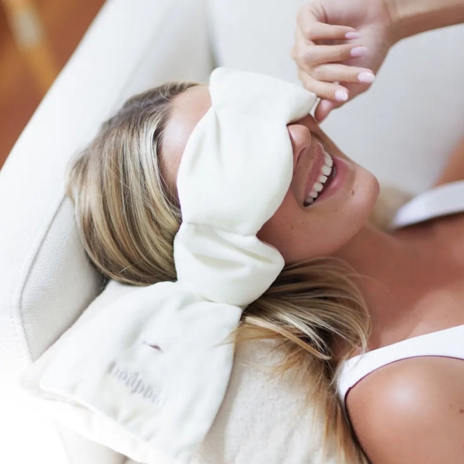 Person reclining on a light-colored couch, wearing a beige Nodpod weighted sleep mask shaped like a bow, smiling.