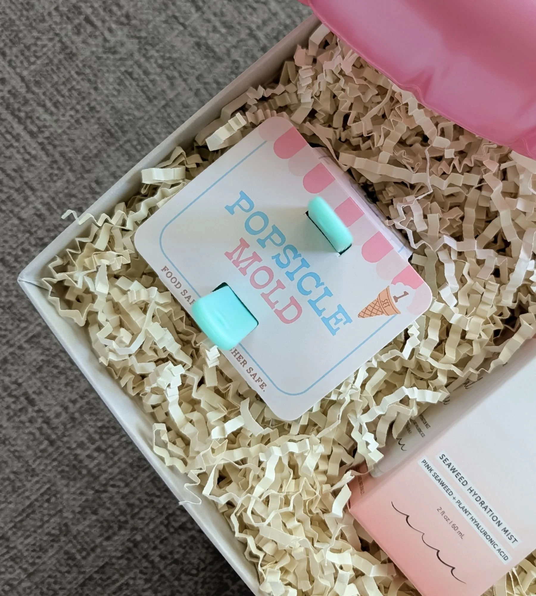 Pool‑themed gift box with a set of two popsicle molds a pink flamingo floatie and hydrating rose facial spray all arranged with colorful summer items and premium filler.