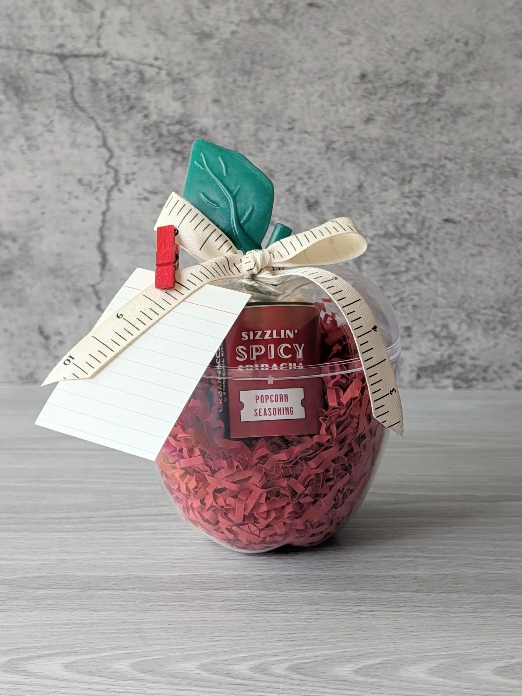 A plastic apple filled with red crinkle paper, a mini popcorn bag, and a small popcorn seasoning, finished with a notebook‑style tag and ruler ribbon