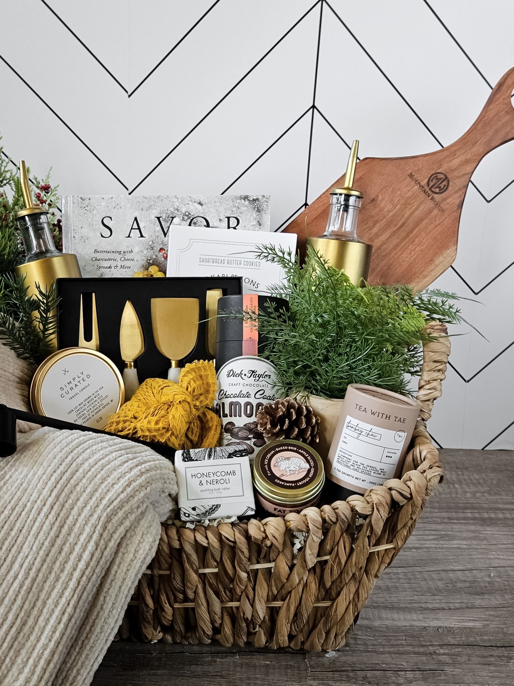 Elegant housewarming gift basket with a cozy throw, cookbook, gourmet treats, bath tablet, candle, and stylish home accessories.