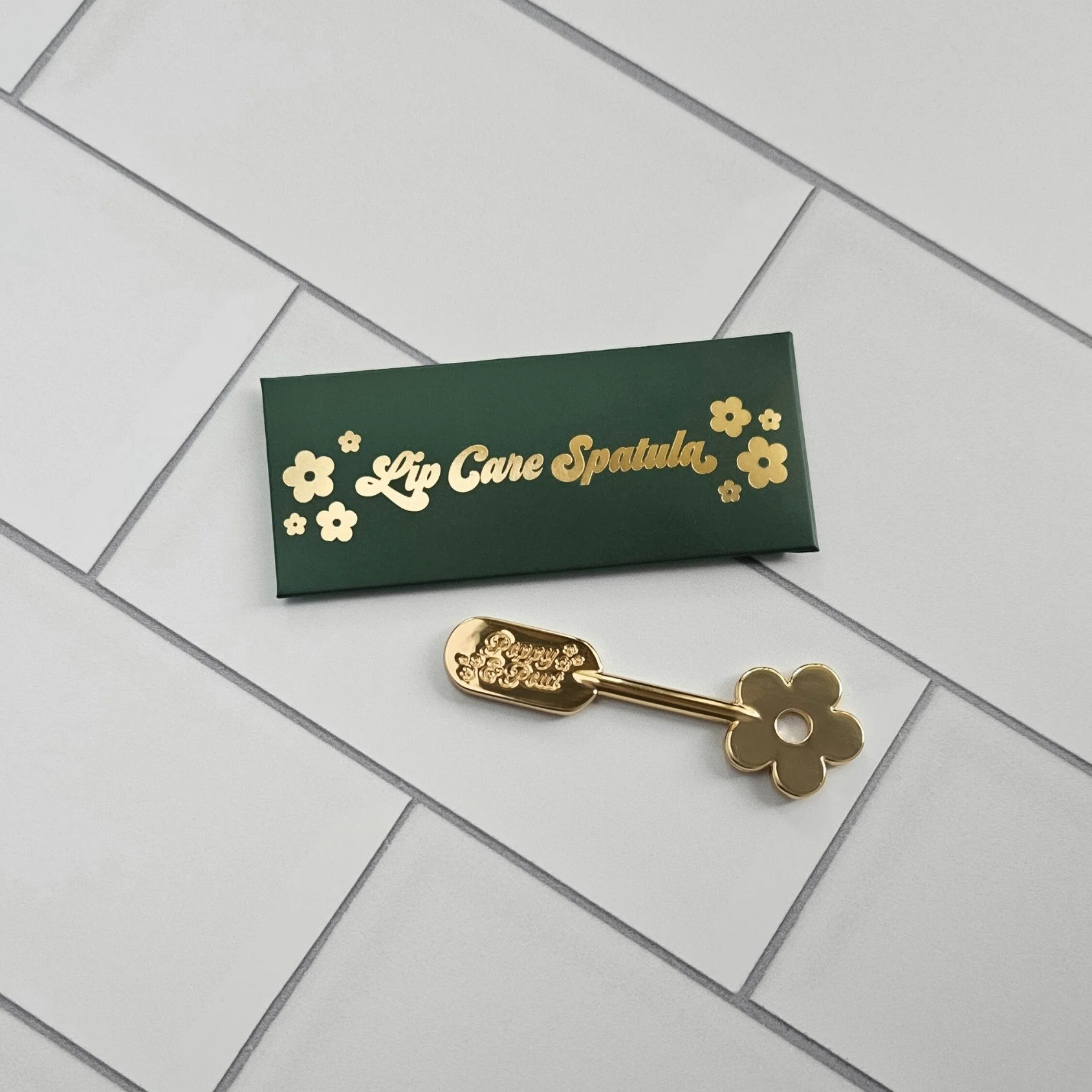 Gold lip scrub spatula by Poppy & Pout with a flower shaped handle, perfect for clean, elegant application and a stylish gift for her.