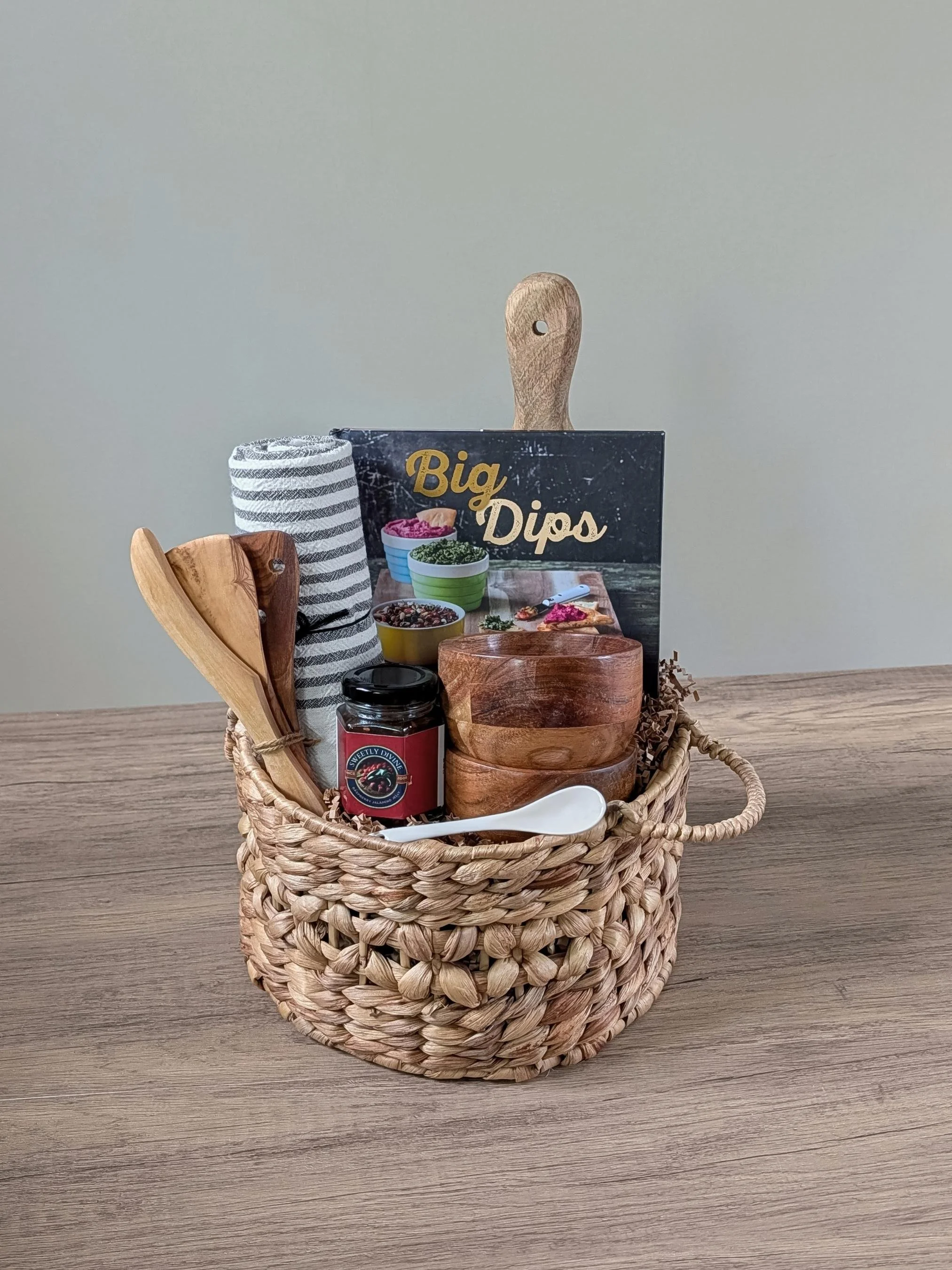 Apéro Bites gift basket with the Big Dips recipe book, striped towel, olive wood trio, dip bowls, serving board, ceramic spoon, and cranberry jalapeño jelly.