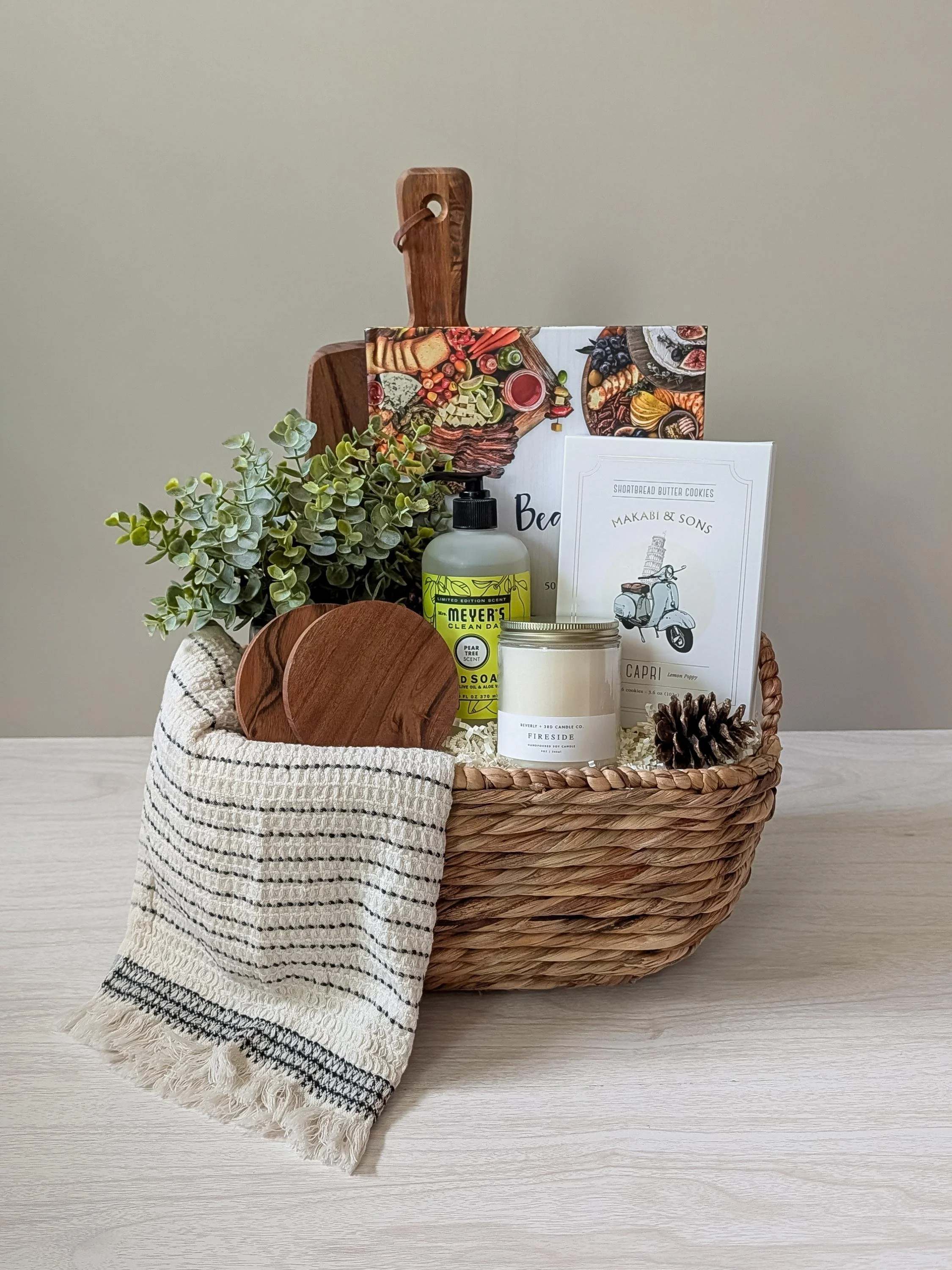 Beautiful housewarming gift basket with a wood board, striped towel, Fireside candle, Meyer’s soap, wooden coasters, pinecone, greenery, and gourmet Capri cookies arranged neatly in a woven basket.