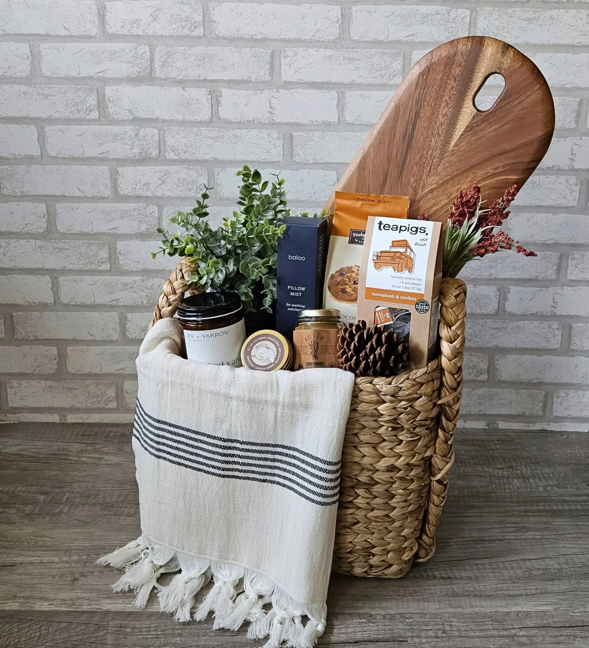 Rustic housewarming gift basket featuring cookie mix, honeybush rooibos tea, smoked vanilla candle, bourbon caramel sauce, Wasatch wildflower honey, pillow spray, a Turkish hand towel, and a large wood serving board.