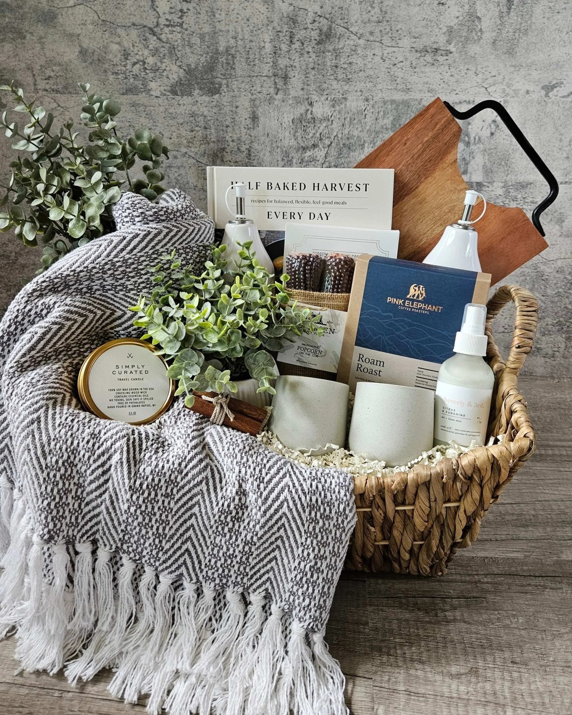 Luxury gift basket featuring a cozy grey and white throw blanket, gourmet coffee, popcorn on the cob, large serving board, Harvest Cookbook, Japanese stone coffee mugs, a candle, and linen spray.