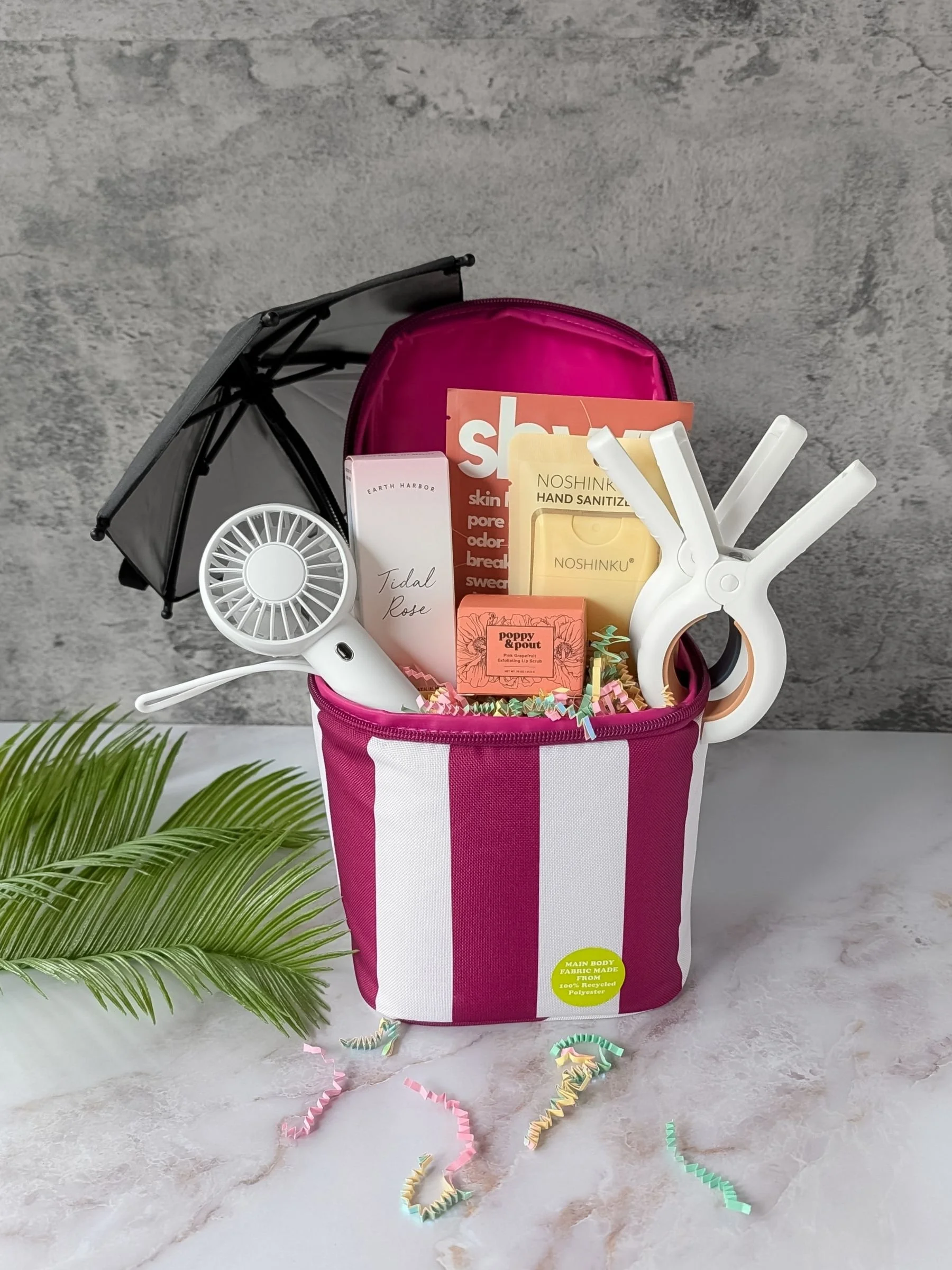 A magenta and white striped drink cooler filled with summer essentials including a portable fan, phone umbrella, facial spray, sanitizer, lip scrub, body wipe, and pool towel clips, arranged with colorful crinkle paper.