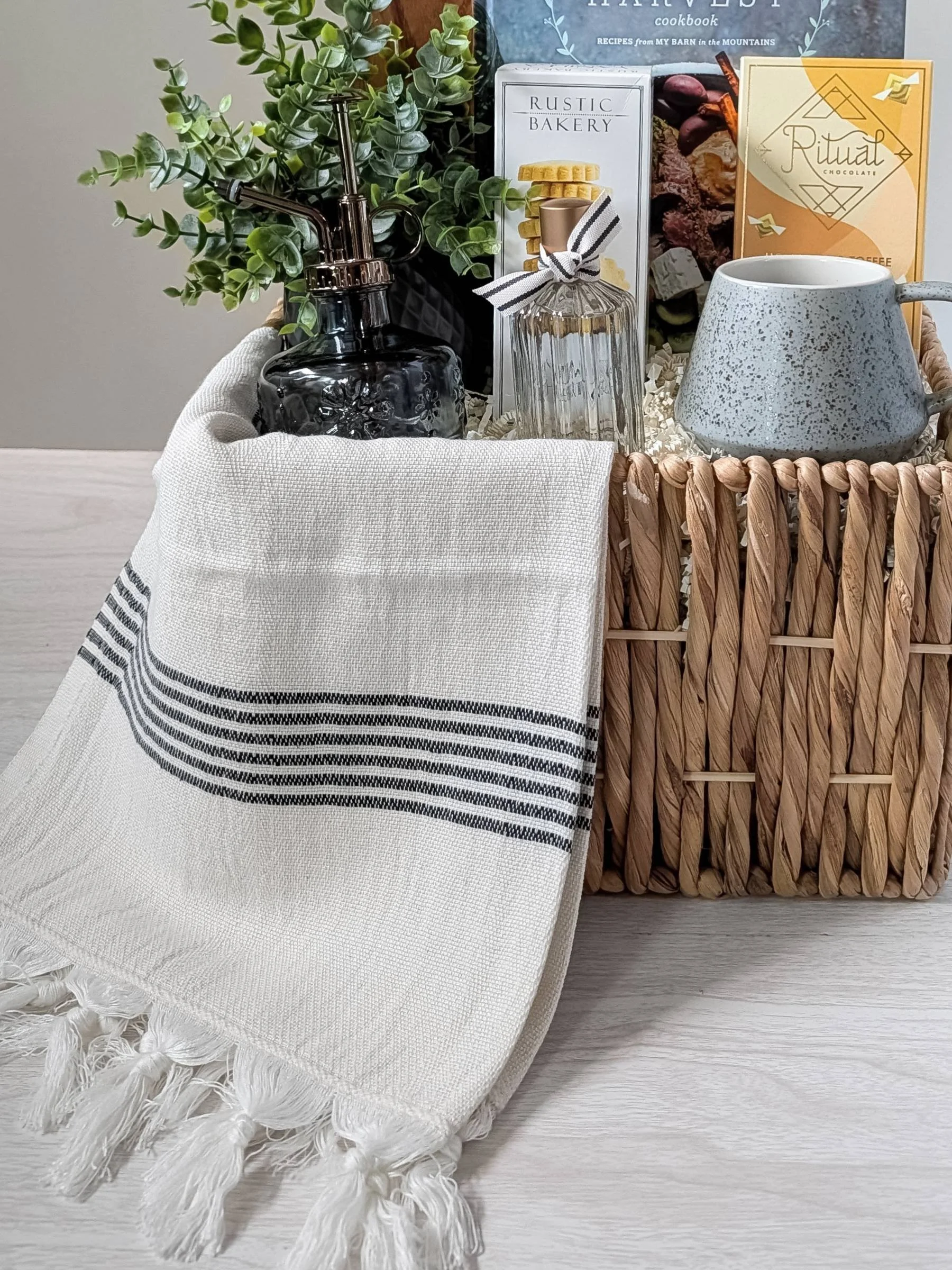 Gourmet housewarming gift basket with cookbook, chocolate, cookies, linen spray, towel, mug, serving board, and home décor.