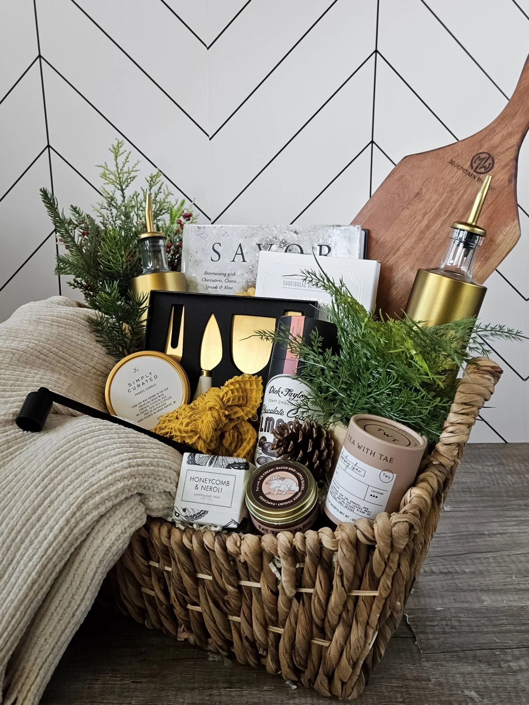 Cozy neutral gift basket with a soft throw, gourmet treats, bath tablet, candle, cookbook, and luxe wood board.