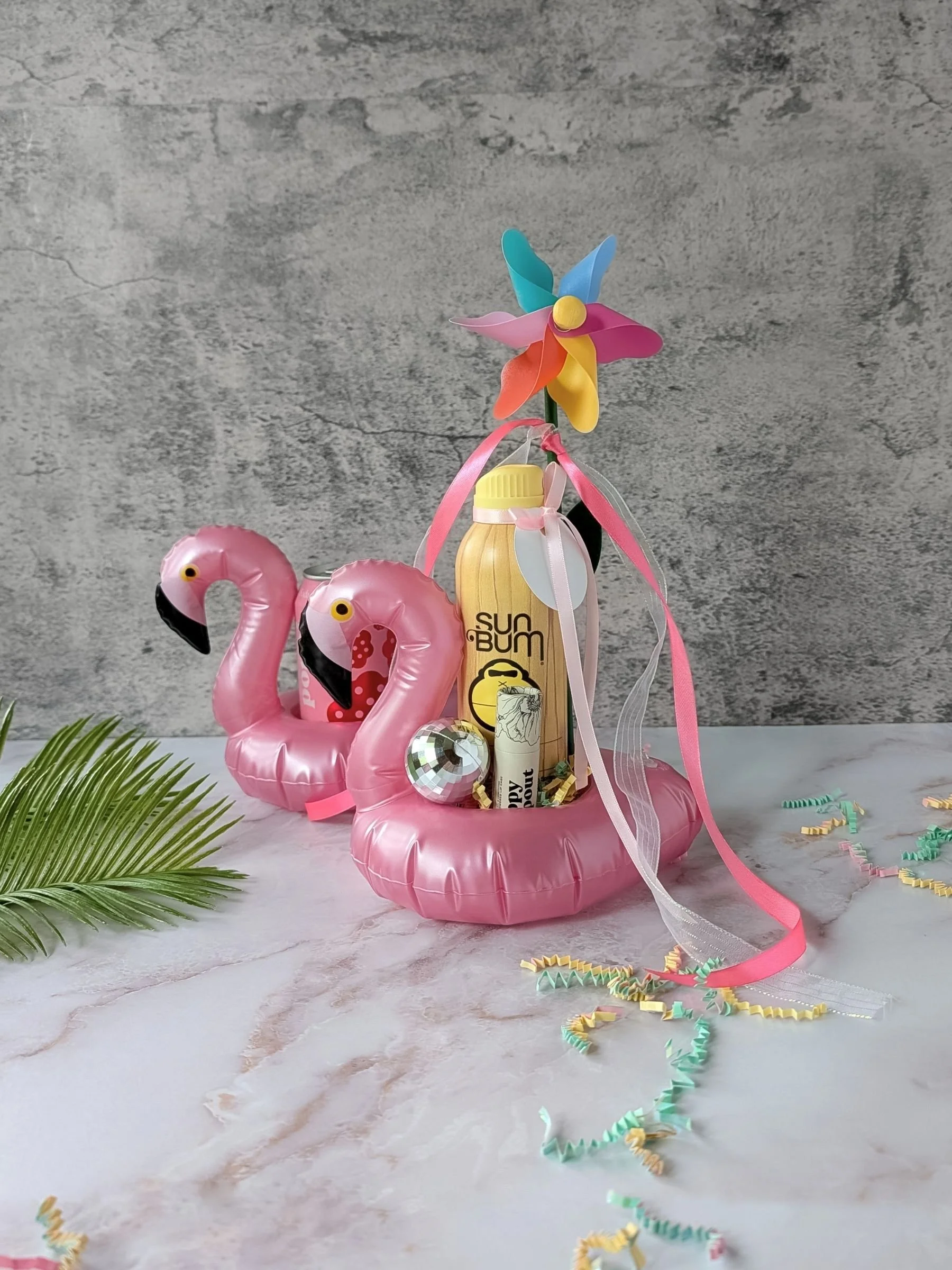 A pink flamingo drink floaty holding a Sun Bum SPF bottle and a Poppy & Pout lip balm, tied with a pink ribbon and gift tag