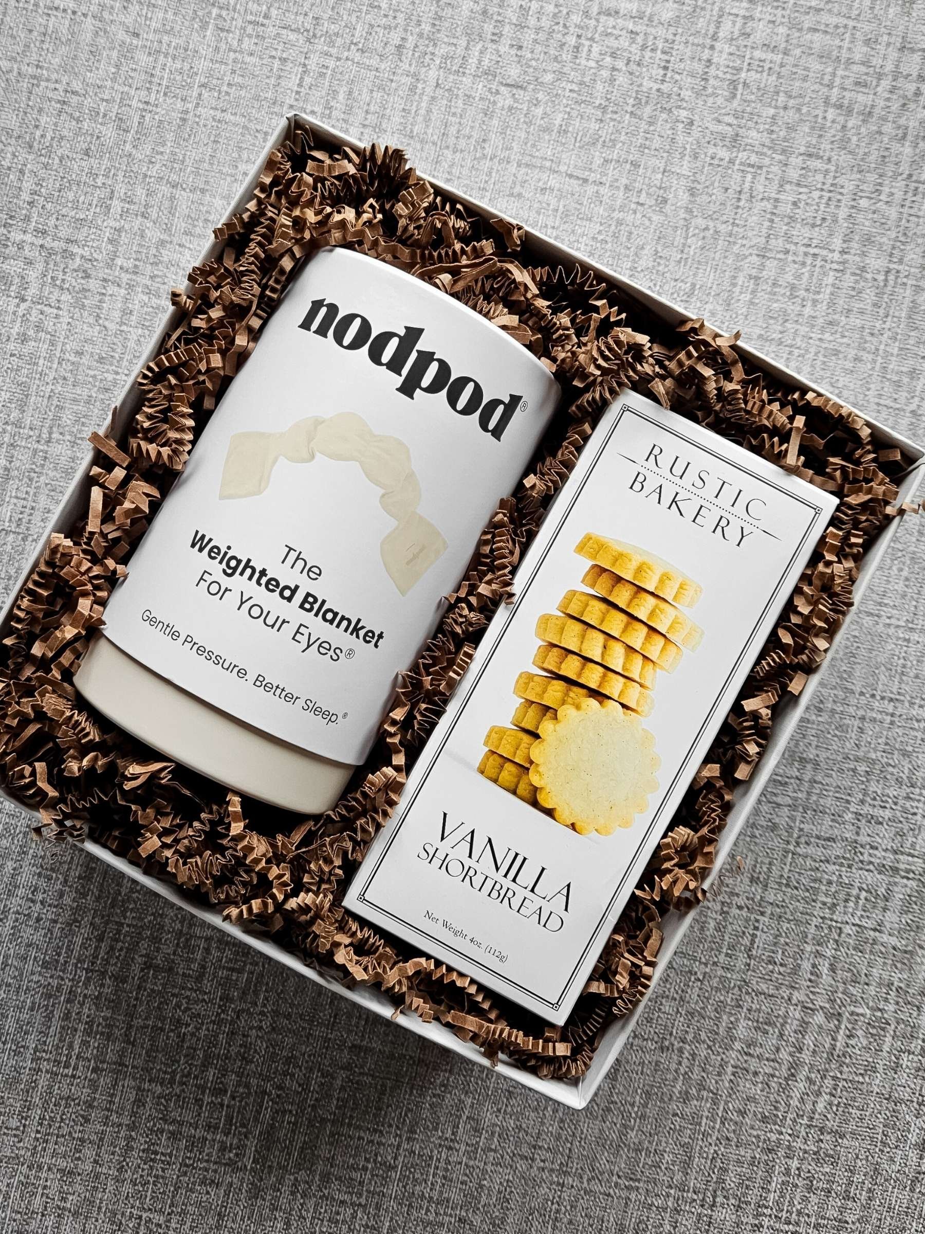 A gift box filled with brown crinkle paper holding a Nodpod weighted sleep mask and a box of Rustic Bakery vanilla shortbread cookies.