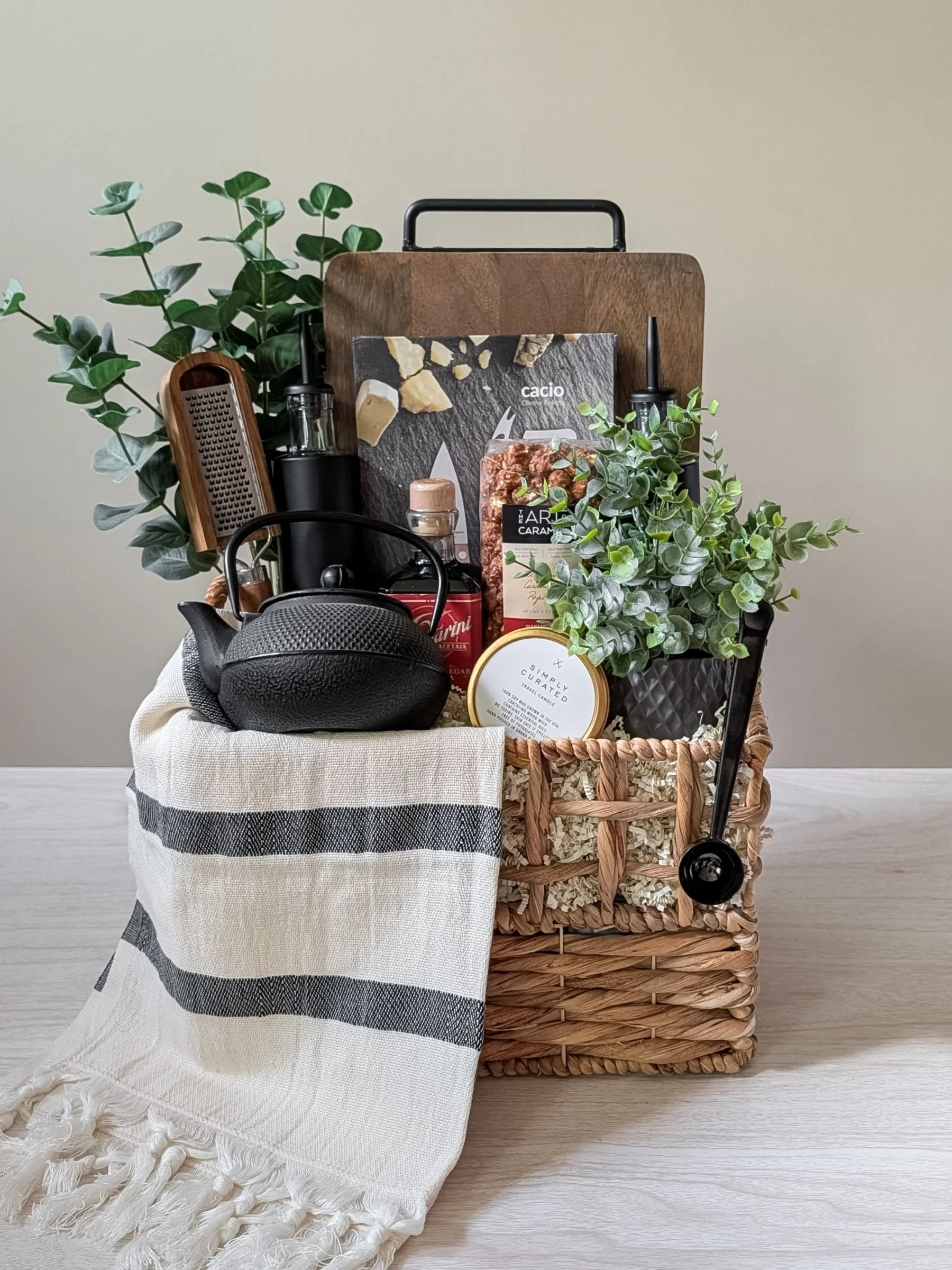 Contemporary housewarming basket styled with a wood board, matte black accents, gourmet popcorn, balsamic vinaigrette, and a striped hand towel.