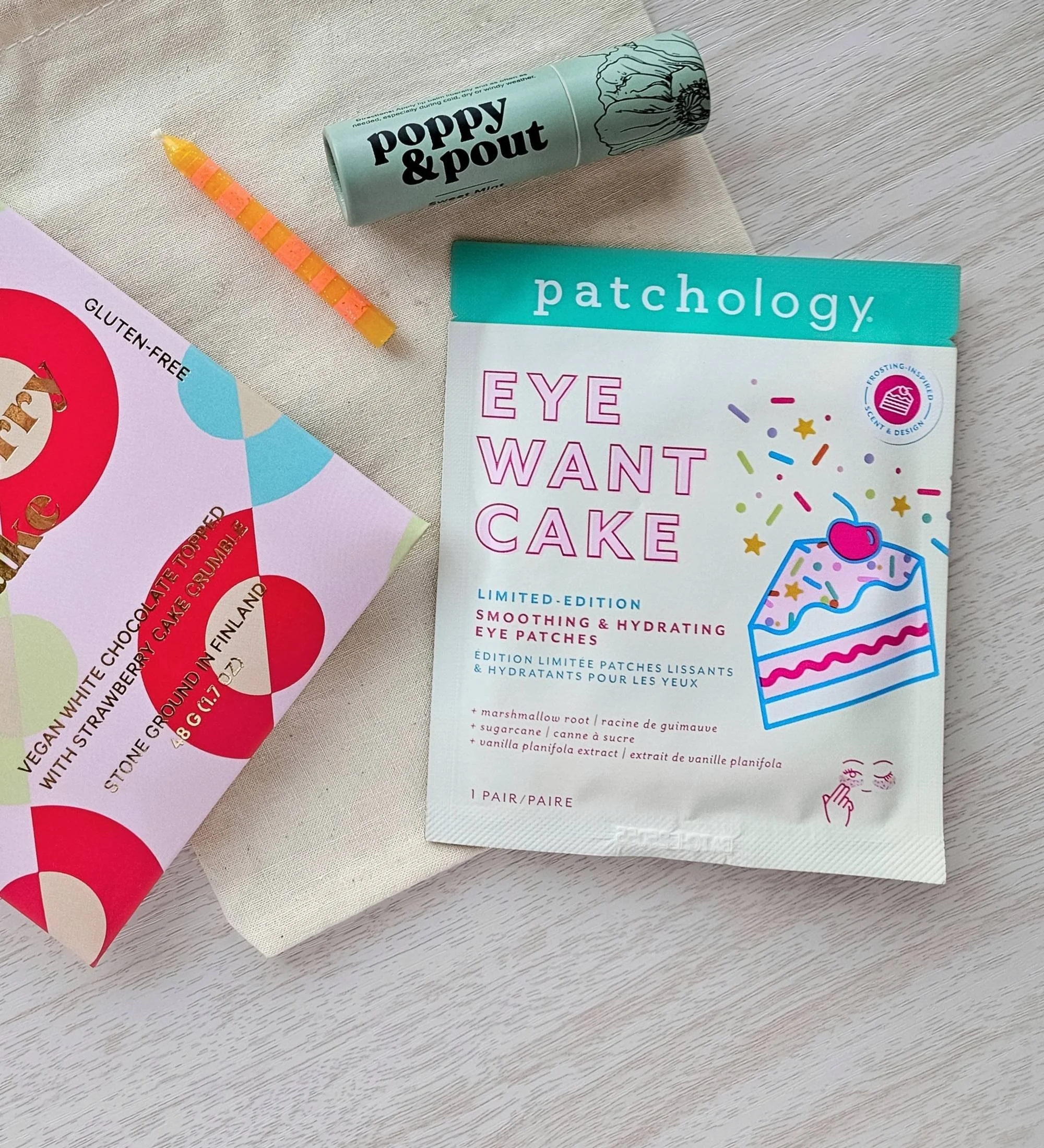 Colorful birthday gift set with candle, Patchology eye gels, Goodio chocolate bar, and Poppy & Pout lip balm styled on a cotton pouch.