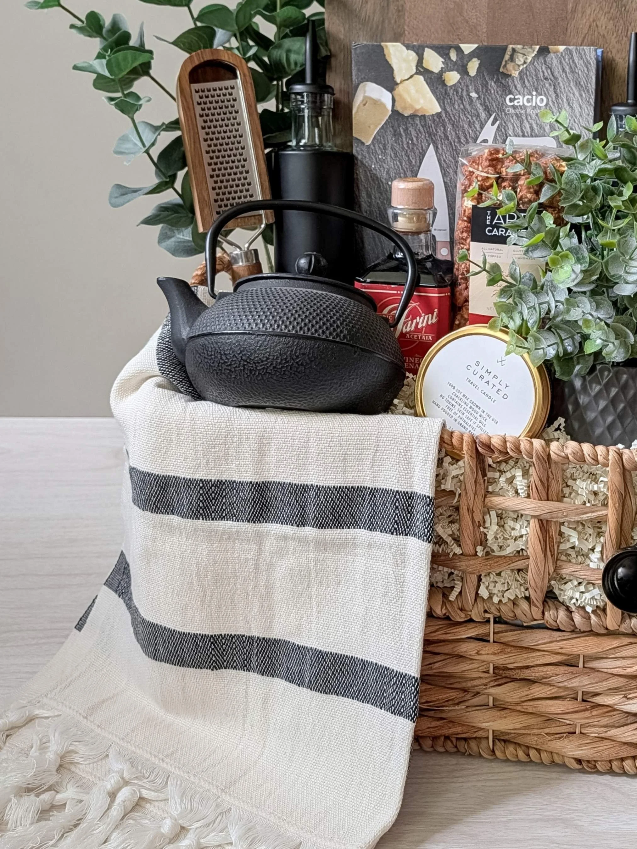 Modern housewarming basket with a large wood serving board, matte black oil and vinegar dispensers, gourmet popcorn, wood grater, and a striped hand towel.