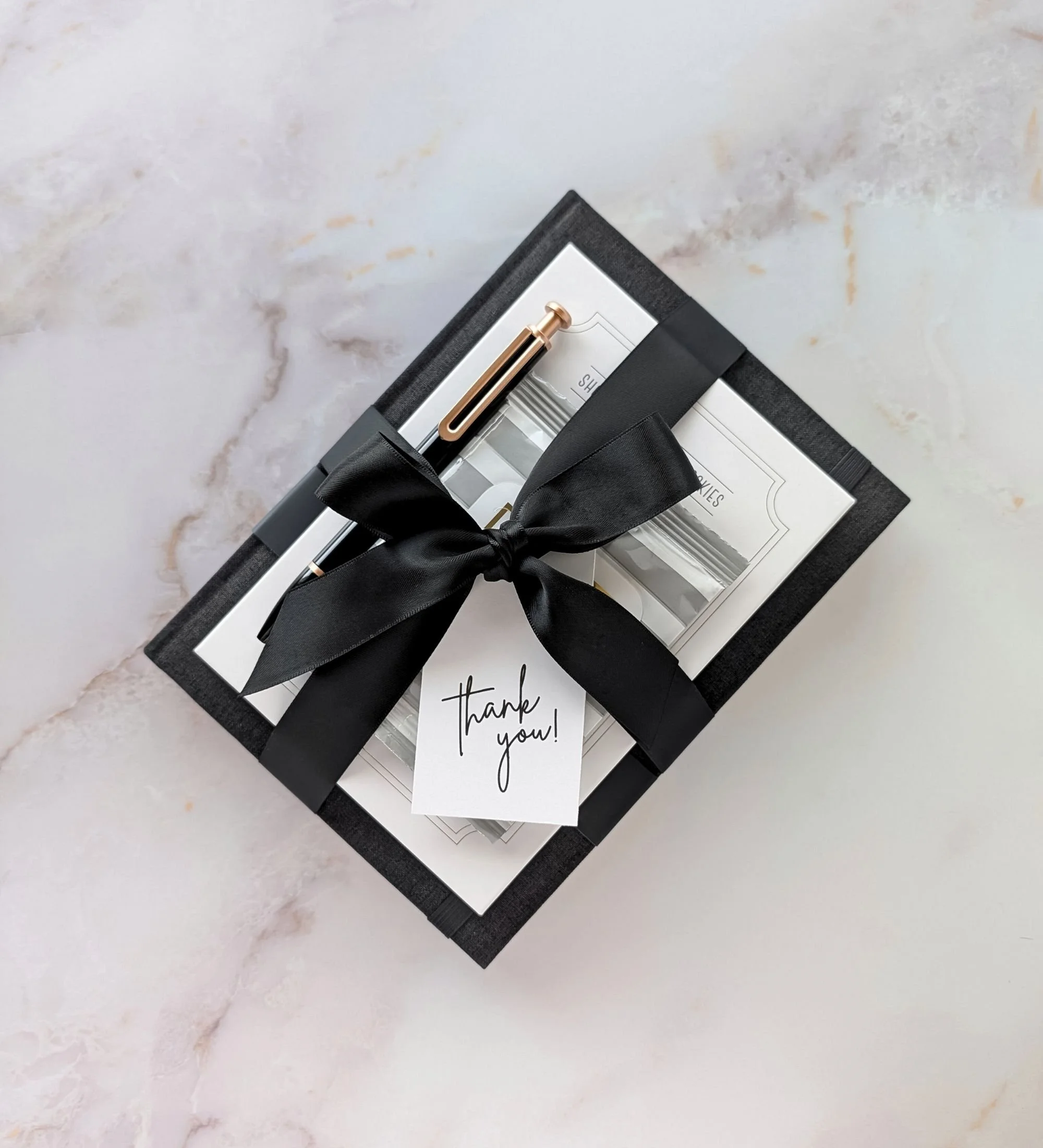 The Ledger modern executive appreciation gift with black notebook, gold pen, cookies, and sleek neutral accessories.